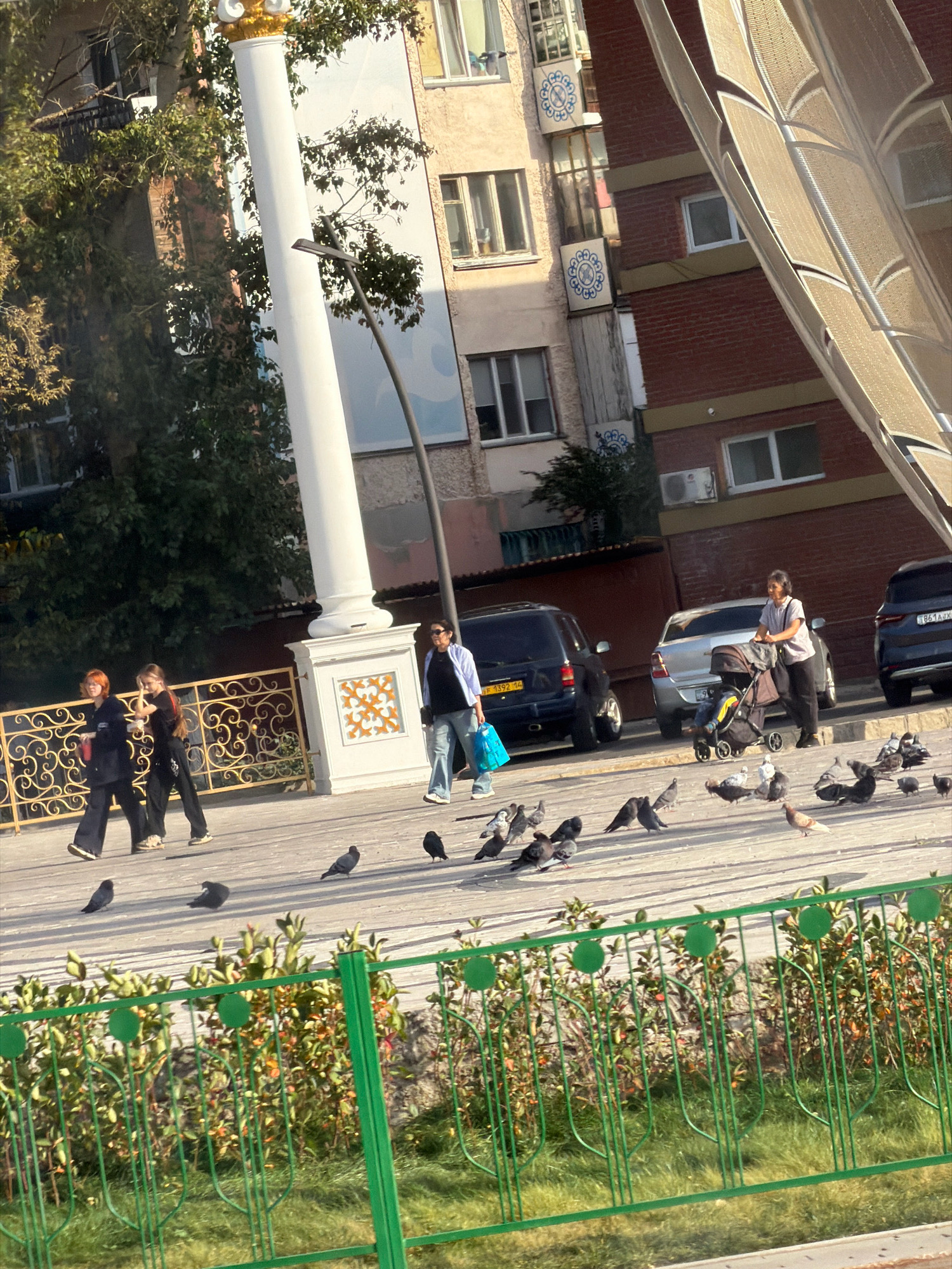 This image depicts a scene on a street in Pavlodar, Kazakhstan, likely in an urban environment. In the foreground, a flock of pigeons are gathered on a paved surface. A green metal fence with a floral motif separates the paved area from a grassy strip with shrubs and trees.
In the mid-ground, several people are walking along a sidewalk or pathway. Two young women with auburn and dark hair are walking together, both wearing dark clothing. Another woman, wearing a blue shirt and jeans, is walking with a stroller. A man is seen in the background near the strollers.
Behind the pedestrians, a row of parked cars are visible, including a dark SUV and a silver sedan. Buildings with multiple stories form the backdrop of the scene. One building has a large white column in the foreground, decorated with an orange and white pattern at its base. Another building is constructed of red brick and has several windows and air conditioning units visible. The overall lighting suggests it is daytime, with sunlight casting shadows.