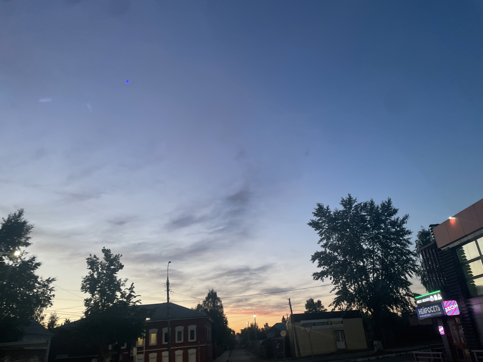This image depicts a street scene in Votkinsk, Russia, captured during dusk or early evening. The sky transitions from a deep blue at the top to a warm orange and pink near the horizon, indicative of sunset, with scattered light grey clouds.
In the foreground and midground, the silhouettes of numerous trees, their leaves dark against the sky, frame the street. Several buildings are visible. On the left, a traditional-looking red building with white-framed windows stands out. A streetlight pole with overhead power lines is positioned towards the center.
On the right side of the image, a more modern building features prominent neon signs. One large sign displays "НЕЙРОСЕТЬ" (NEUROSET) and below it, "ПЕЧАТНЫЙ САЛОН И КОПИЦЕНТР" (PRINT SALON AND COPY CENTER). Another illuminated sign reads "OZON," indicating a pickup point for the Russian e-commerce platform. Above the "NEUROSET" sign, smaller text mentions "МЯСО / МОЛОКО" (MEAT / MILK) with accompanying icons, suggesting a food or convenience store. Further down the street on the right, a yellow building is partially visible, with text that is difficult to fully decipher but appears to be "СУДОВАЯ МЕБЕЛЬ".
The street itself appears quiet, and no people are visible, contributing to a peaceful atmosphere. A small, bright blue dot, likely a lens flare or sensor artifact, is noticeable in the upper left portion of the sky.