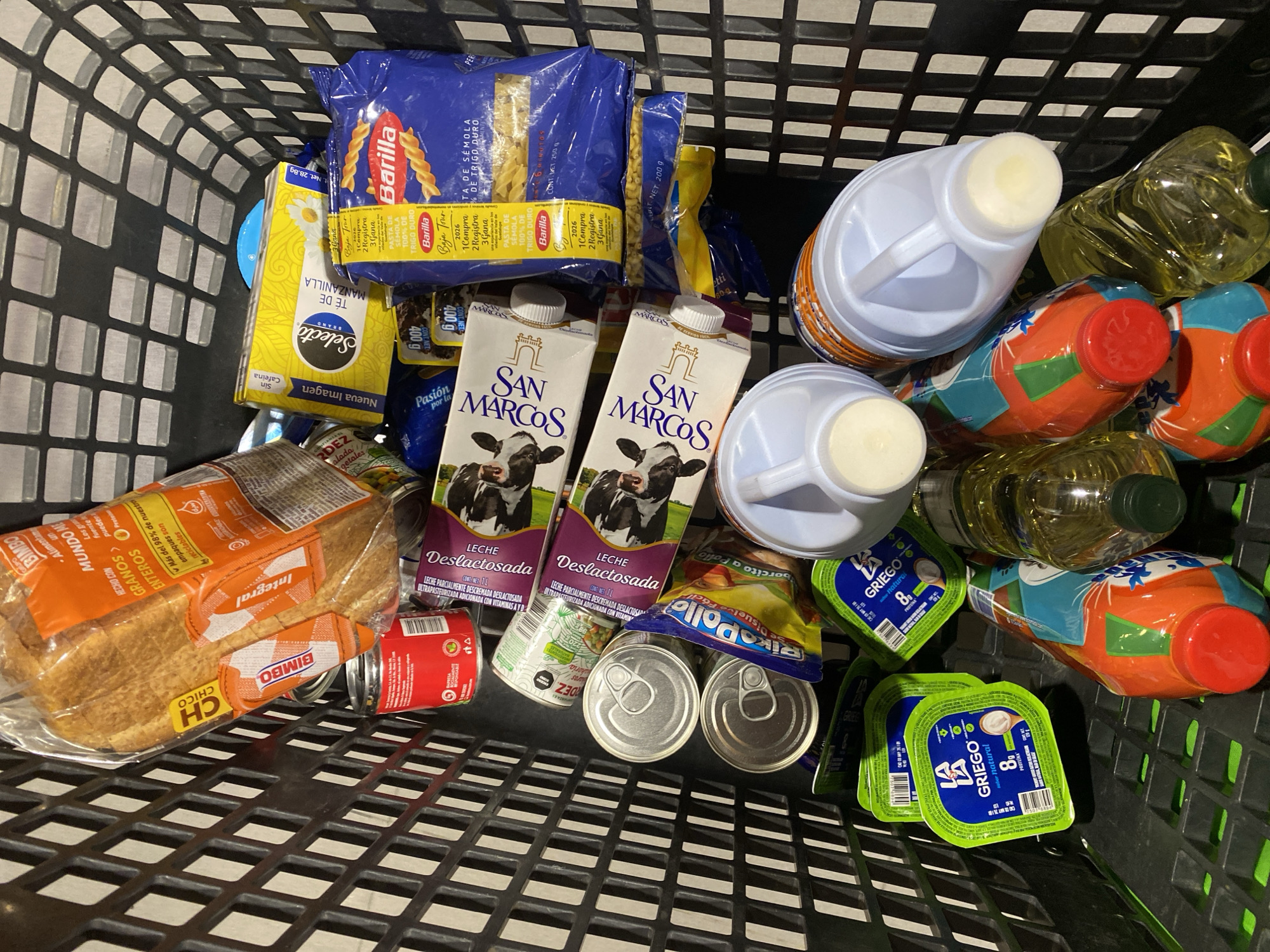 A shopping cart is filled with various grocery items. These include two cartons of San Marcos Lactose-Free Milk, a loaf of Bimbo whole wheat bread, cans of tuna and vegetables, pasta (Barilla), Selecto Manzanilla tea, yogurt (LA Griego), vegetable oil, and juice containers. The setting is a supermarket in San Bernardino Tlaxcalancingo, Mexico, as evidenced by the brand of milk and other products. There are no people visible in the image.