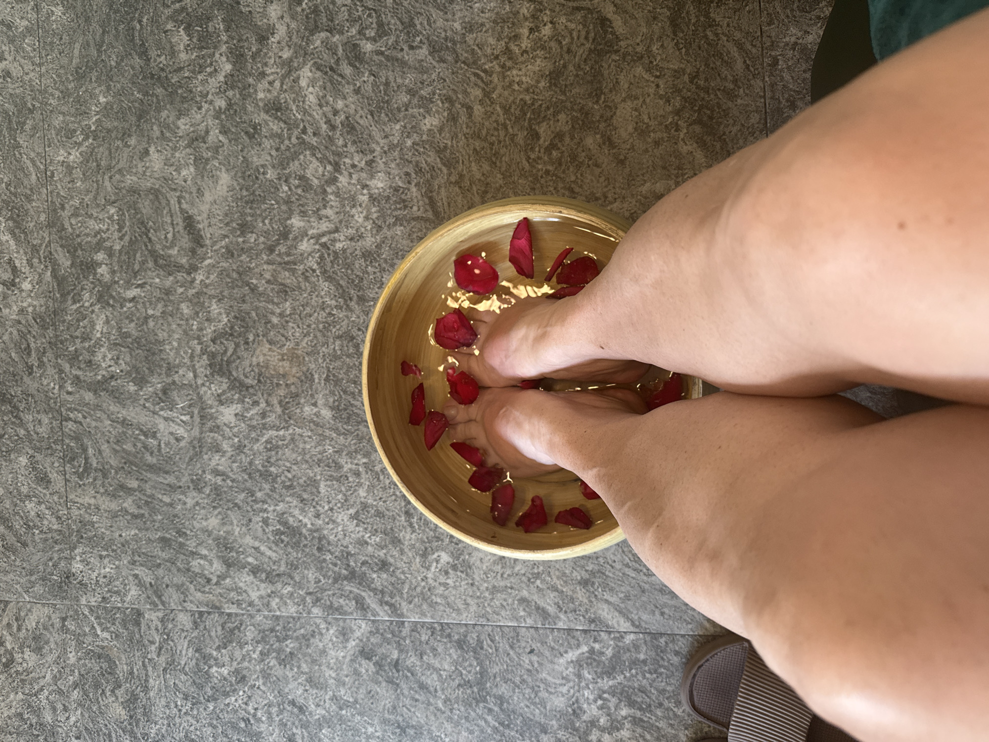 The image shows a top-down view of a person's legs and feet soaking in a shallow wooden bowl filled with water and scattered red flower petals. The person is seated, with the bowl placed on a gray tiled floor. A partially visible slipper rests on the floor near the person's leg. The context suggests the action of a foot soak, potentially in a spa or domestic setting. The image's location is unknown.