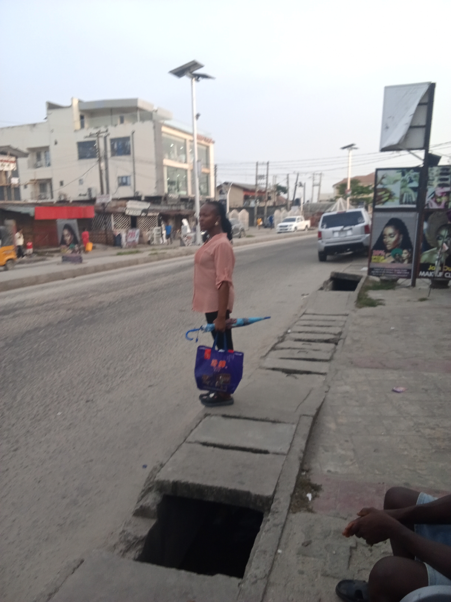 This outdoor shot captures a bustling urban street scene in Ado, Nigeria. The central subject is a woman of African descent, standing on a concrete sidewalk, looking towards her left with a slight smile. She wears a light peach-colored collared shirt with rolled-up sleeves and dark trousers, holding a blue umbrella in her left hand and a blue and purple patterned tote bag in her right.
The sidewalk features large, uneven concrete slabs covering a drainage system, with one section exposing a dark, open ditch. In the bottom right foreground, parts of another person, likely male, are visible, seated on the ground in shorts and sandals.
Across the asphalt road, which has a narrow median strip, vehicles are present, including a white SUV and a white sedan further down. The background is dominated by commercial buildings, mostly light-colored, some with multiple stories and glass facades. A red sign is visible below one building on the left. Streetlights equipped with solar panels punctuate the scene.
On the right side of the road, a large billboard structure displays advertisements. One prominent poster features a woman with dark skin and makeup, suggesting a beauty-related product or service. Partial text "MAKE UP CL" is visible on one of the signs, likely part of "MAKE UP CLASS" or similar. The sky is overcast or hazy, indicating either early morning or late afternoon lighting. The overall scene depicts a typical commercial area in a Nigerian city.