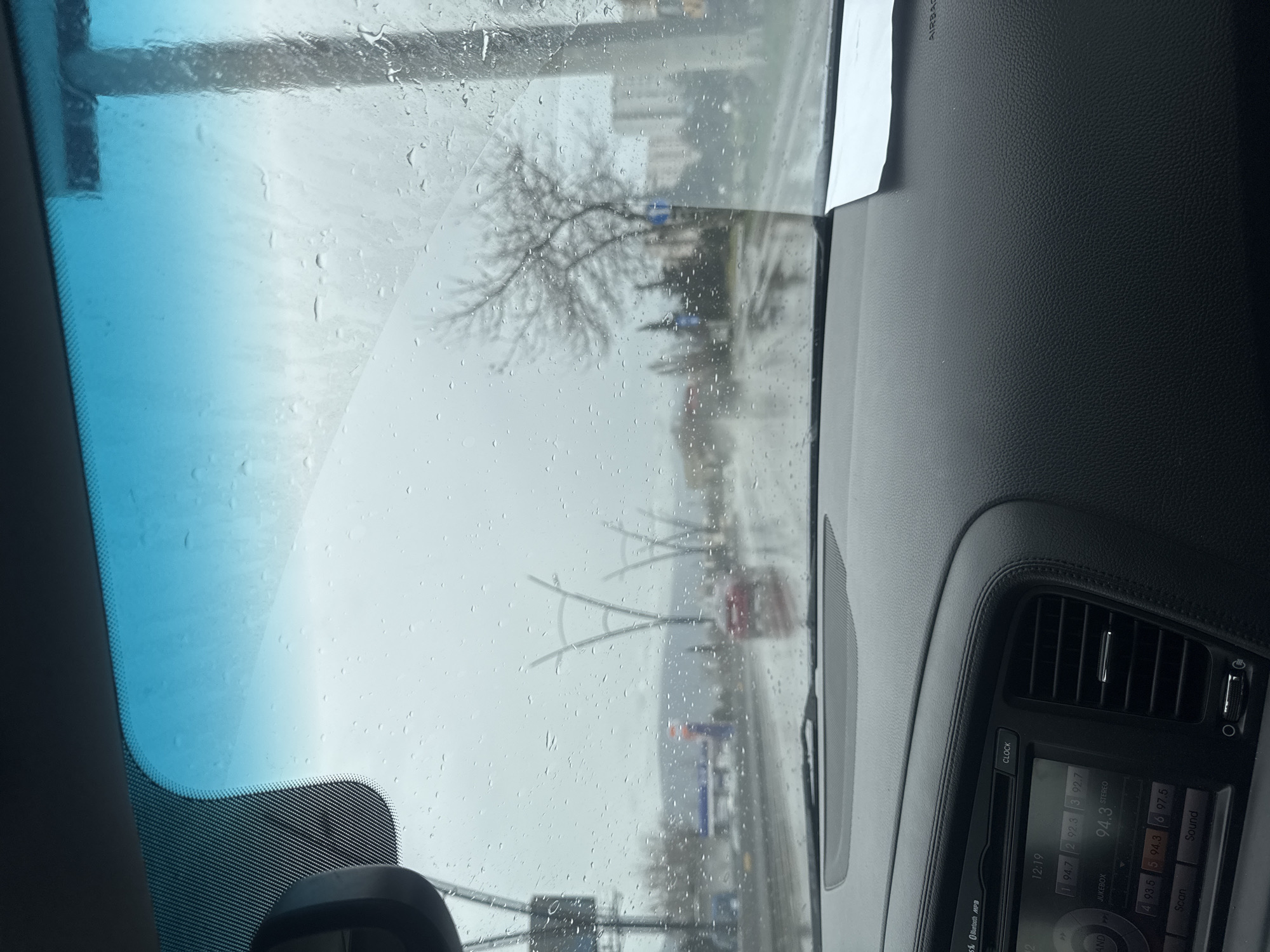 The image depicts an interior view from a vehicle looking out through a rain-splattered windshield on an overcast day in Tbilisi, Georgia. The car's dark, textured dashboard occupies the lower frame, featuring an infotainment system displaying "12:19" and "94.3 STEREO," along with "Bluetooth MP3" and radio presets. A white paper is positioned on the passenger-side dashboard, which includes "AIRBAG" text.
The exterior scene shows a wet, multi-lane asphalt road reflecting light. A red vehicle is visible ahead in the distance. The road is lined with distinctive, modern, V-shaped streetlights, identifying the location as a major artery such as Vazha-Pshavela Avenue in the Saburtalo district. A blurred gas station with blue and red elements appears on the left. Sparse, bare trees and multi-story buildings are visible on either side of the road, with hills forming the distant horizon under a gray sky. No individuals are clearly discernible.