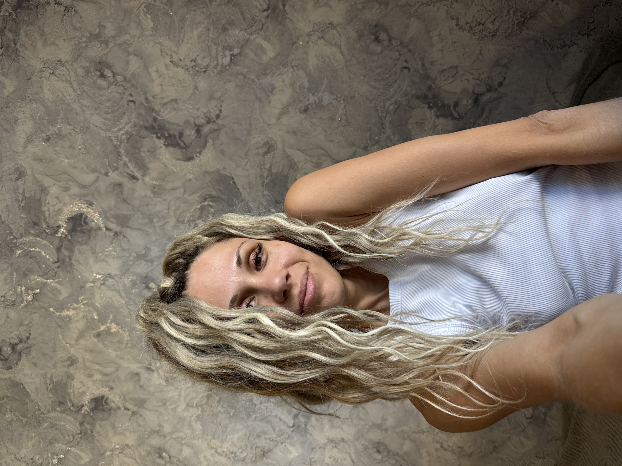 A woman with blonde, curly hair, wearing a white tank top, looks at the camera with a slight smile. Her right arm is bent and resting on her chest. She is positioned in front of a textured, grey wall.