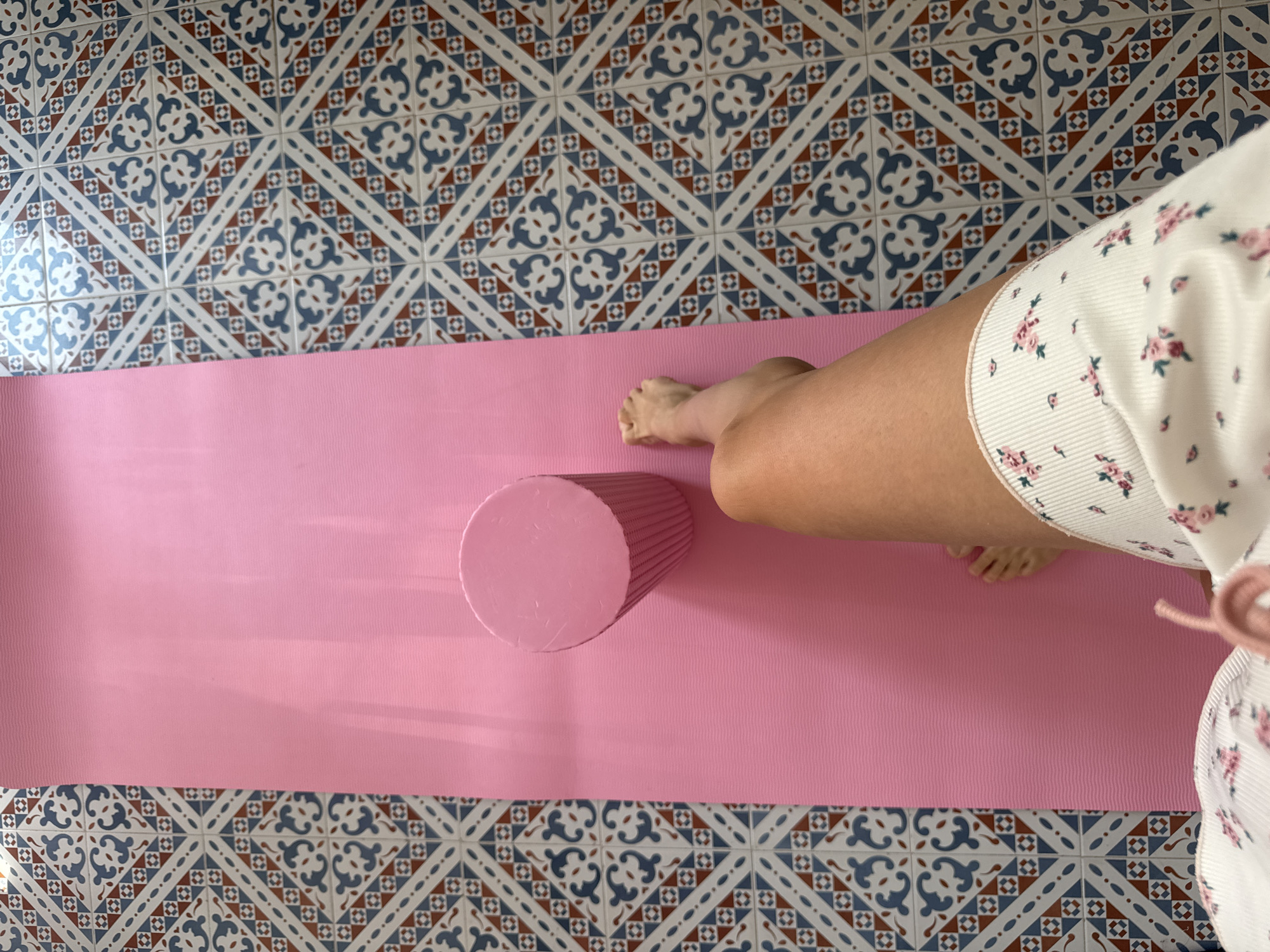 An overhead shot captures a pink yoga mat laid out on a floor with intricate, patterned tiles. The tiles feature a repeating geometric design in blue, white, and rust colors. A person's bare right leg, from the knee down to the foot, rests on the yoga mat. The individual is wearing white shorts adorned with a delicate floral pattern of pink roses and green leaves. To the left of the leg, a pink cylindrical foam roller stands upright on the mat. The scene appears to be indoors, possibly a home gym or a dedicated wellness space, given the presence of the yoga mat and foam roller, and the decorative tiling. The location is specified as Isola delle Femmine, Italy.