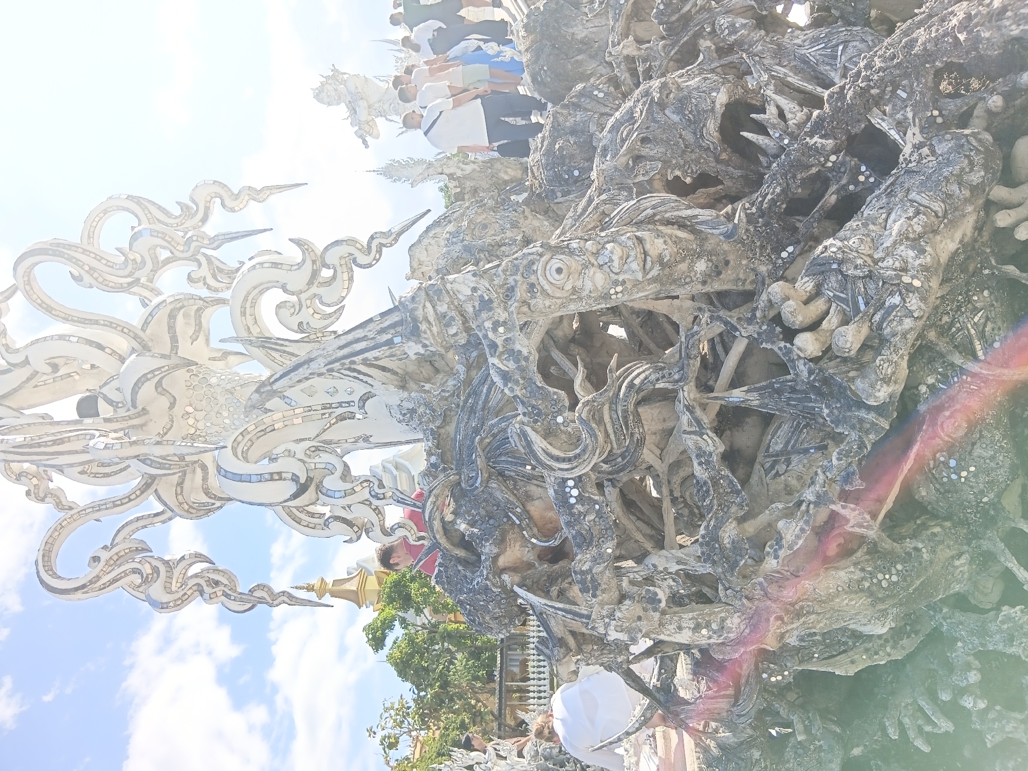 The image depicts an outdoor scene in Ban Rong Khun, Thailand, featuring a large, intricate sculpture predominantly made of white material adorned with reflective fragments, likely glass or mirrors. The sculpture is detailed with organic and monstrous forms, including visible faces with eyes and mouths, skeletal structures, and swirling, serpentine elements. In the foreground, the sculpture is dense and appears to be constructed from a chaotic arrangement of intertwined forms.
Several people are present in the scene. In the middle ground, a man in a red shirt is visible, looking towards the sculpture. To the left, a person in a white shirt is also looking at the sculpture. Further back, a group of individuals, including men and women dressed in casual clothing, are standing and observing the artwork. The environment is bright and sunny, with a blue sky and scattered clouds. Green foliage and other architectural elements, including a golden spire, are visible in the background, suggesting a temple or cultural site. Lens flare is present in the lower left and center of the image, indicating strong light sources.
