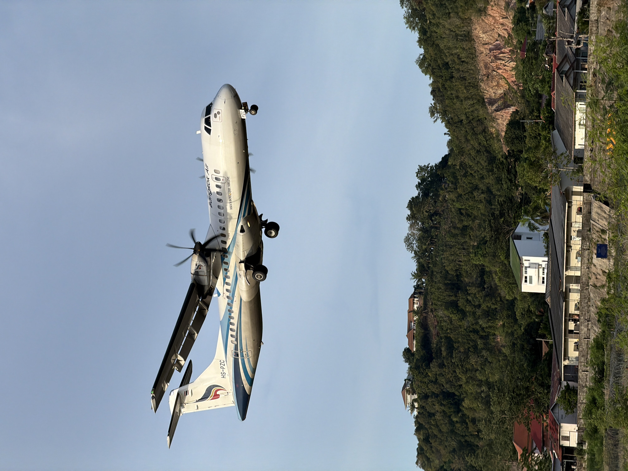 An ATR 72-500 aircraft, identified by the registration HS-PZC and the livery of Bangkok Airways, is captured mid-flight against a clear blue sky. The airplane is a turboprop model with two propellers visible, one in motion. The airline's branding, "Bangkok Air," and the tagline "ASIA'S BOUTIQUE AIRLINE" are clearly visible on the fuselage. The aircraft appears to be either descending or ascending, with its landing gear deployed.
Below the aircraft, a verdant, hilly landscape unfolds, characteristic of Baan Bo Phut, Thailand. The hills are densely covered with lush green trees. Nestled on the slopes of the hills and in the valley below are several buildings. Some of these appear to be houses with red and white roofs, while others are more modern structures, including a white building with a green roof. The foreground features a more rustic setting with low-lying buildings and vegetation, suggesting a residential or semi-rural area. A steep, rocky cliff face is visible on the right side of the frame, adding to the natural topography of the area. The overall scene depicts a serene, tropical environment with a modern aircraft traversing the sky above it.