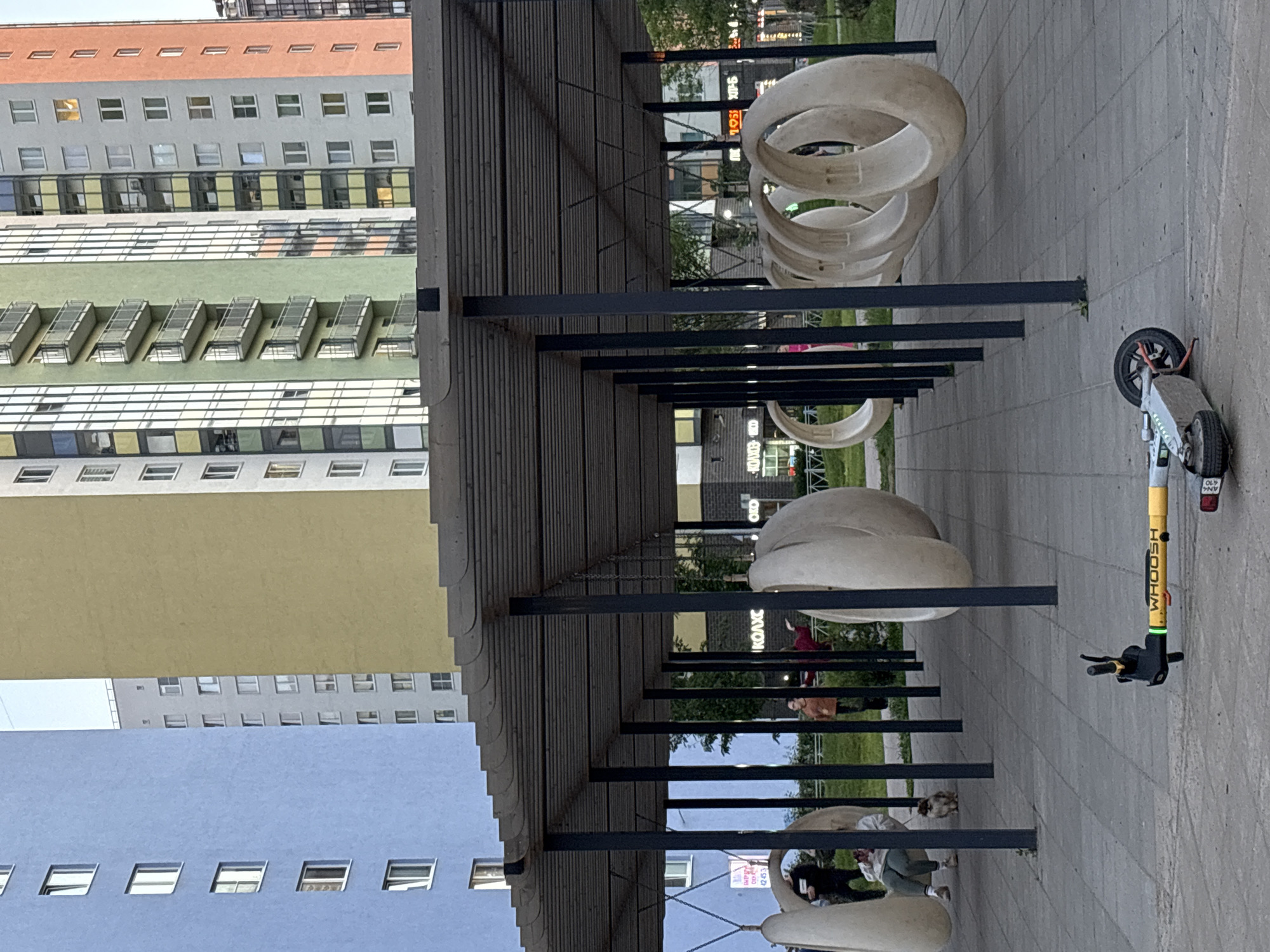 In the city of Unknown, Russia, an outdoor scene unfolds beneath a modern, slatted canopy. The canopy is supported by dark vertical poles, creating a shaded area over a paved walkway. A yellow and black electric scooter lies on its side on the pavement in the foreground, its handlebars pointing left. Behind the canopy, a row of large, white, ring-shaped sculptures are suspended from the ceiling by chains, some appearing as if they are intended for seating or play.
In the middle ground, several people are visible under the canopy. One individual, wearing a white top and green pants, sits on one of the ring sculptures with their legs extended. A small dog sits near them on the pavement. Further back, another person stands in an orange shirt, and another in a dark top is partially visible. In the background, a row of colorful, multi-story apartment buildings stands out. The buildings are painted in various shades, including light blue, yellow, green, gray, and coral. Windows are visible on each floor of the buildings. The overall environment suggests a public space, possibly a residential courtyard or park, with elements of modern architecture and recreational features.