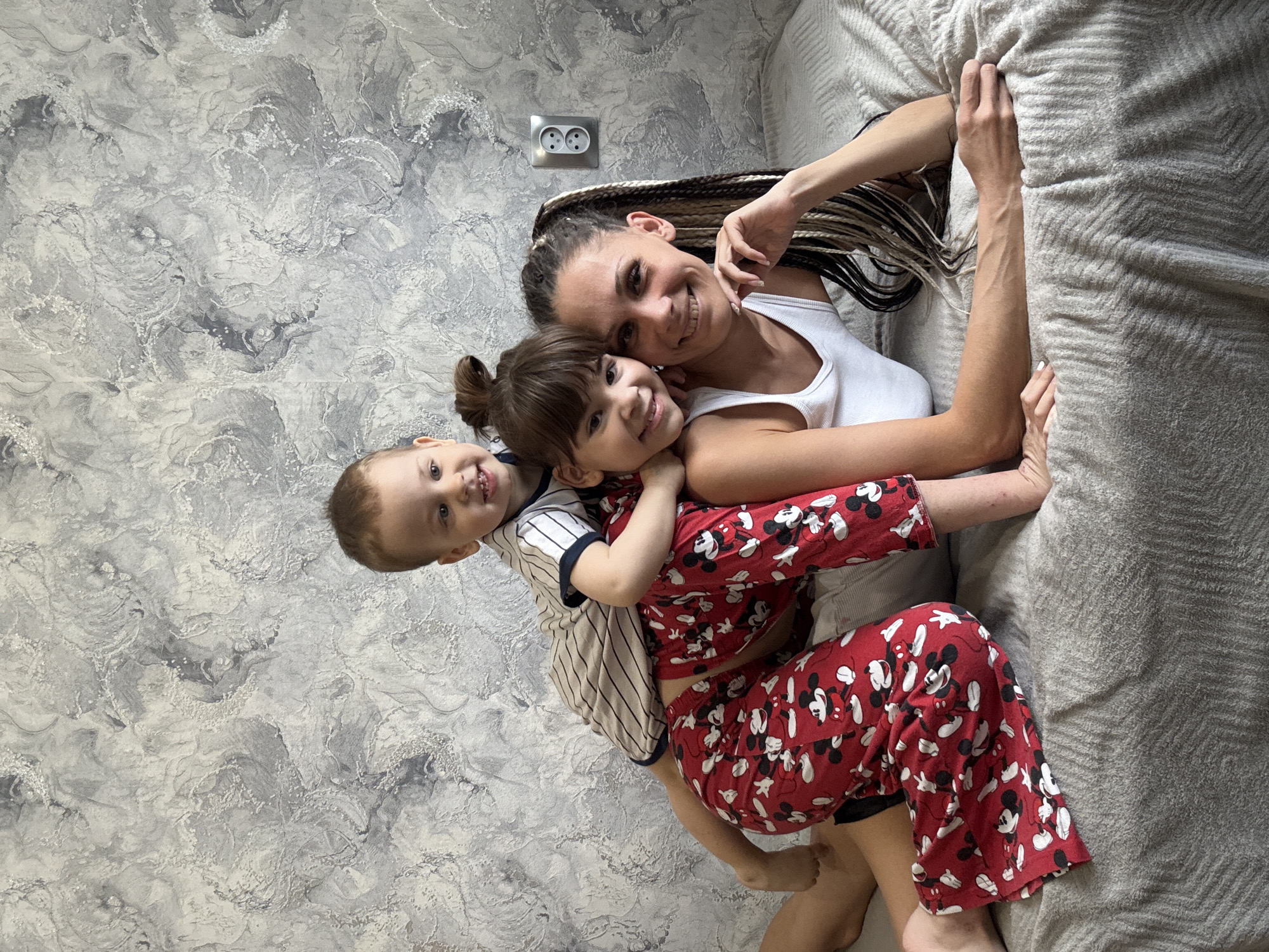 An overhead shot depicts a woman and two children (a boy and a girl) lying closely together, likely on a bed or soft surface, in an indoor setting. All three subjects are looking towards the camera with warm smiles, conveying a sense of happiness and affection.
The woman, positioned on the right side of the frame, has long, multi-toned braided hair that extends behind her. She wears a white tank top and is embracing the young girl who is nestled against her. The girl has light brown hair tied in a small bun and is wearing bright red pajamas adorned with Mickey Mouse characters. To the left of the girl, the young boy is visible, also smiling. He wears a striped short-sleeved shirt.
The background consists of a textured wall with a grey and white marbled or swirled patterned wallpaper. An electrical outlet with two sockets is visible on the wall above the woman's head. On the right side of the image, a light grey, textured blanket or bedspread is visible, suggesting a bed or sofa. The scene appears to be set in a home in Rusanovo, Russia. There is no visible text in the image.