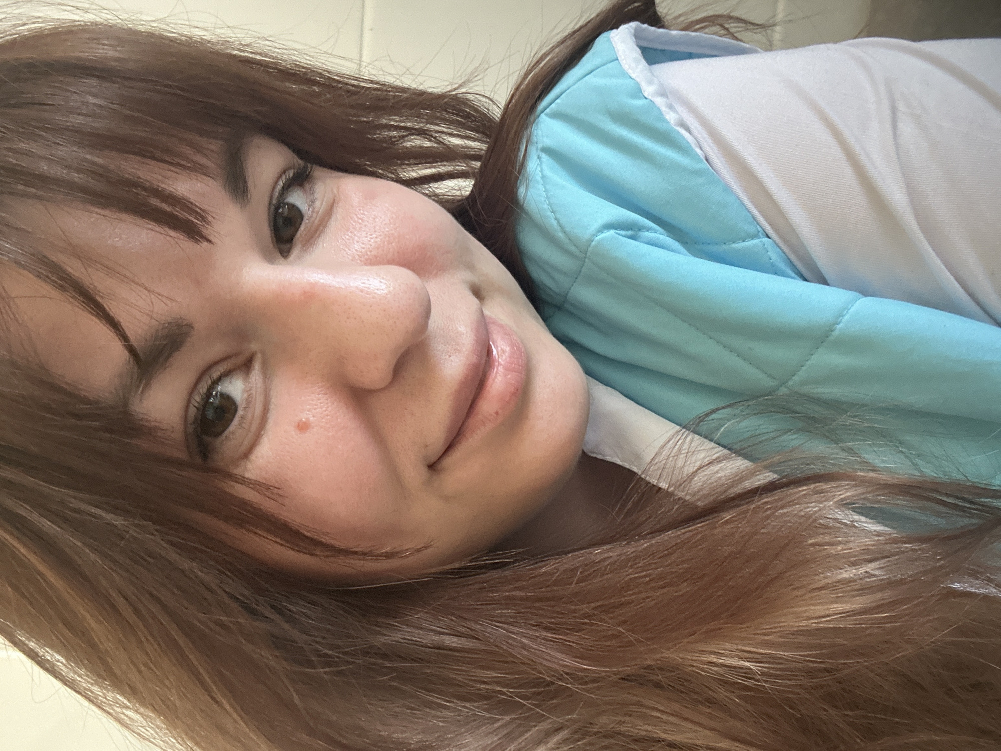 A close-up portrait shot depicts a female individual in an indoor setting. The image, purportedly taken in the city of Фоки, Russia (location not discernible from image content), focuses on the subject's face and upper torso. The woman has long, reddish-brown hair with bangs, partially obscuring her right shoulder. Her eyes are dark brown, and she exhibits a subtle, closed-mouth smile. A small, reddish lesion is visible on her left cheek, below the eye. She is wearing a light blue, quilted sleeveless vest over a white long-sleeved shirt. The background is a plain, light-colored surface, possibly a wall, providing no additional environmental details. The lighting is even, suggesting an intentional portrait or selfie.