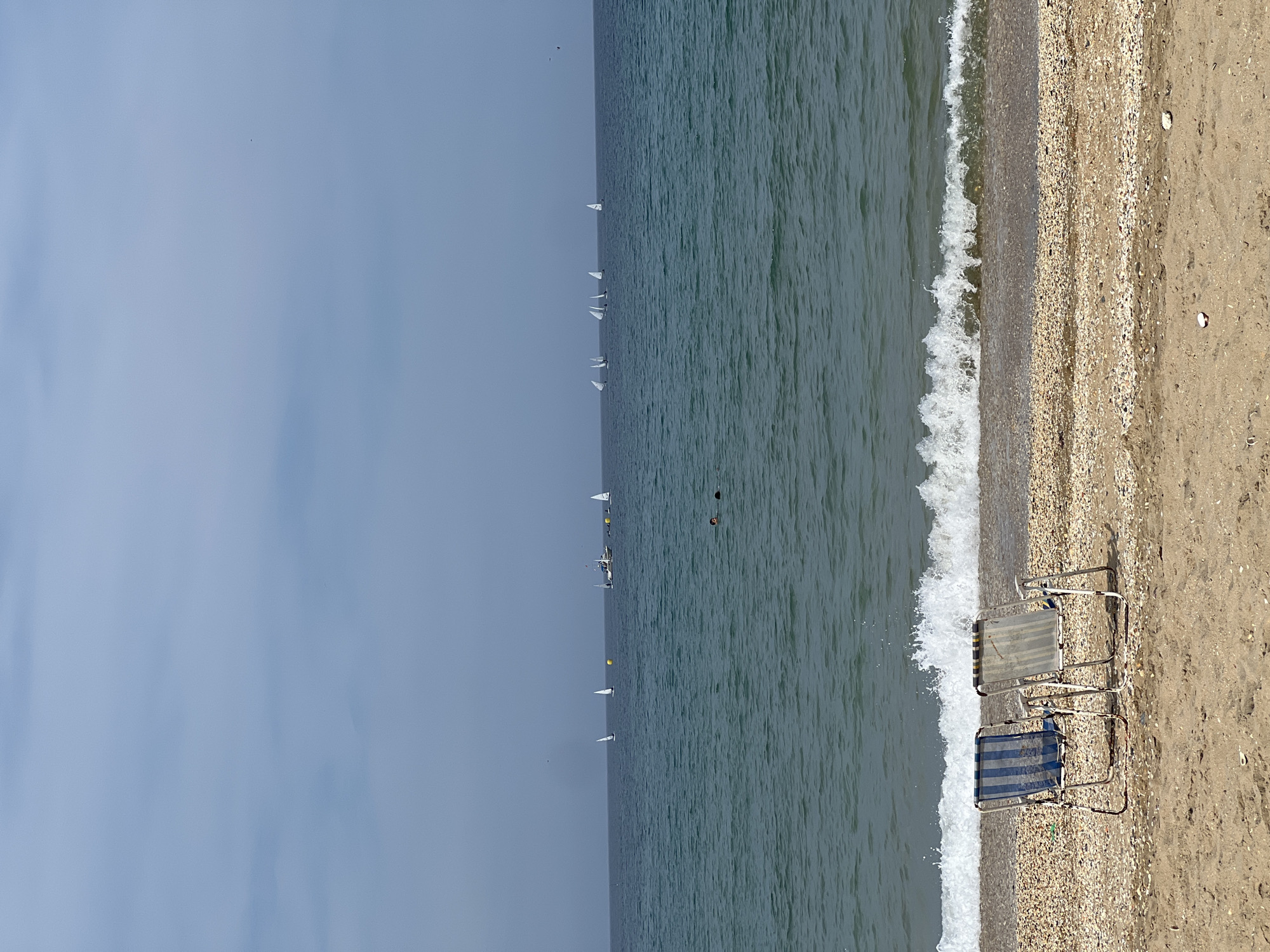 The image depicts a beach scene in M'Diq ⵎⴹⵢⴰⵇ المضيق, Morocco. Two folded beach chairs sit on the sand near the shoreline. The water is visible, with small waves breaking onto the beach. In the distance, there are several sailboats on the water. The sky is overcast. The beach is composed of sand and pebbles.