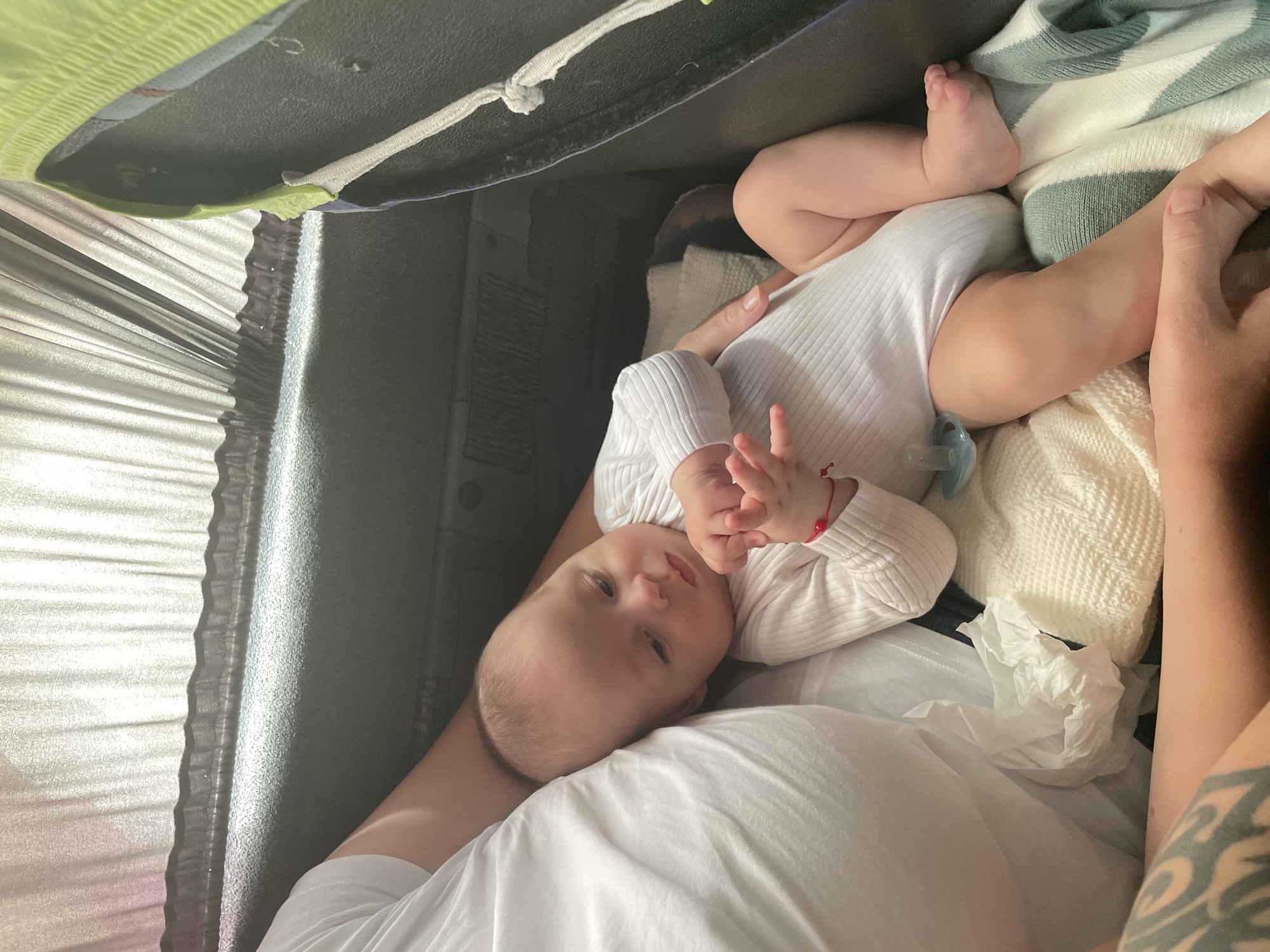A newborn baby is lying on a blanket, held by an adult whose arm is visible on the left side of the image. The baby is wearing a white bodysuit and has a pacifier. The adult has a tattoo on their arm. The scene appears to be indoors, possibly in a vehicle given the presence of a dark-colored seat and window coverings in the background. The image quality suggests it was taken with a mobile device. There is no specific information in the image to indicate the location is in Răuțel, Moldova.