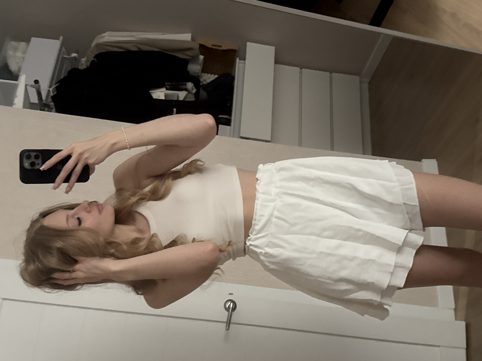 A young woman with blonde, wavy hair is taking a selfie in a dimly lit room, possibly her bedroom. She is wearing a white crop top and white ruffled shorts. Her left arm is raised, holding a black smartphone, and her right hand is in her hair. In the background, a closet is visible with clothes hanging and shelves containing storage boxes. A white dresser with multiple drawers is also present. The floor appears to be made of wood. The scene does not provide any specific indicators to determine its location in the city of Perm, Russia.