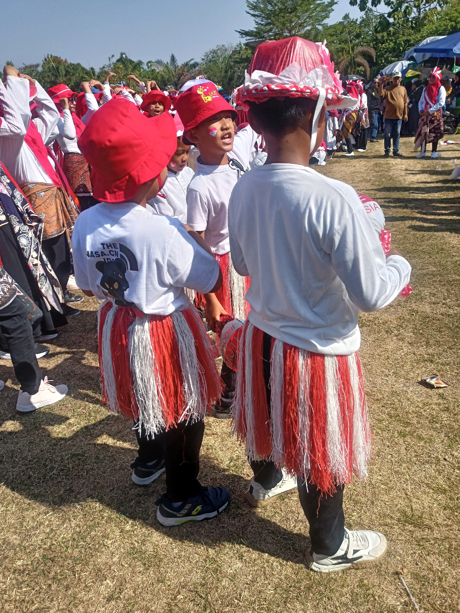 This image captures a vibrant outdoor scene, likely a cultural event or parade, in Sumberlawang, Indonesia. The foreground is dominated by two young people wearing traditional attire. The person on the left has a red conical hat and a white t-shirt with a graphic on the back. Their skirt is a striking red and white fringed skirt, worn over black leggings. The person on the right wears a similarly styled red and white fringed skirt over black leggings, and a white long-sleeved shirt. Their hat is a more elaborate, circular creation adorned with red and white ribbons and what appears to be a plastic covering. Both are facing away from the viewer, engaged in an activity that involves interacting with a crowd.
The background is filled with a diverse group of people, many also dressed in red and white, suggesting a patriotic or celebratory theme. Some are wearing red hats similar to those in the foreground, while others have different headwear or are bareheaded. In the distance, tents and trees are visible, indicating an open field environment. The ground is covered with dry grass. The sky is clear and bright blue, suggesting a sunny day. The overall atmosphere appears to be one of lively participation and community gathering.