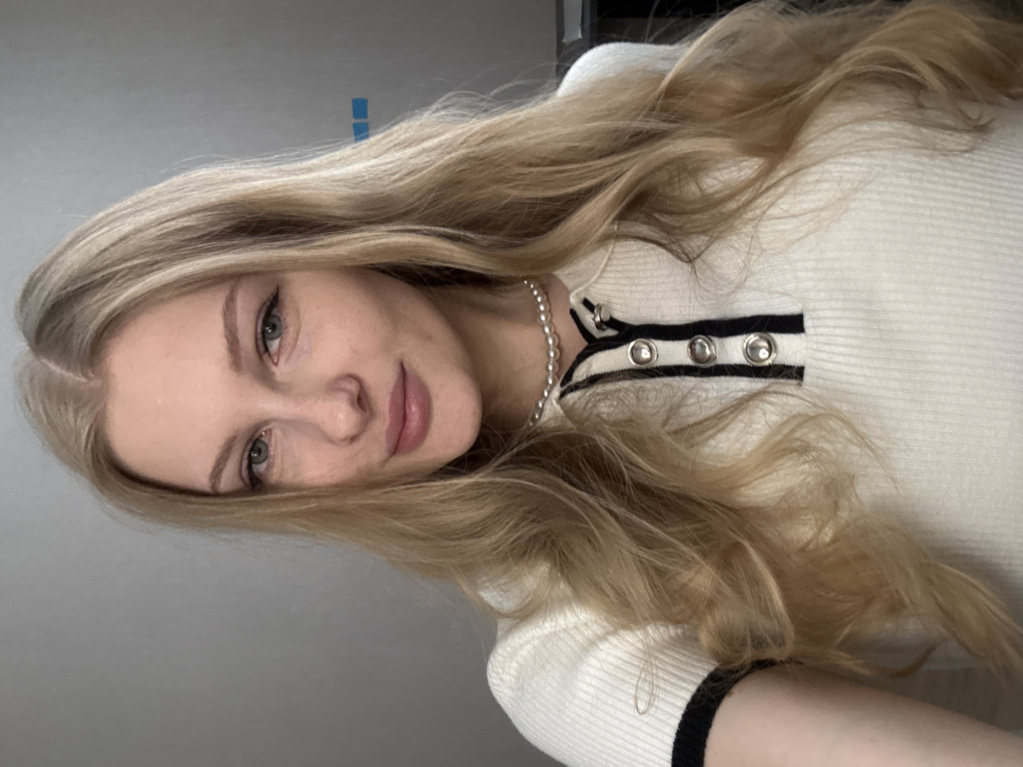 The scene depicts an individual located in the city of Perm, Russia. A young Caucasian woman with long, wavy blonde hair, parted on her left, looks directly at the camera. Her eyes appear to be a light green or blue hue, and she exhibits a slight smile. She is wearing a white, ribbed knit top with a polo-style collar, featuring black trim on the collar, placket, and possibly the sleeve cuffs. The placket contains three silver-rimmed buttons. A pearl choker necklace is visible around her neck. The background is a plain, light grey wall, with two small, rectangular blue markings or objects visible in the upper right quadrant. The lighting is soft and even.