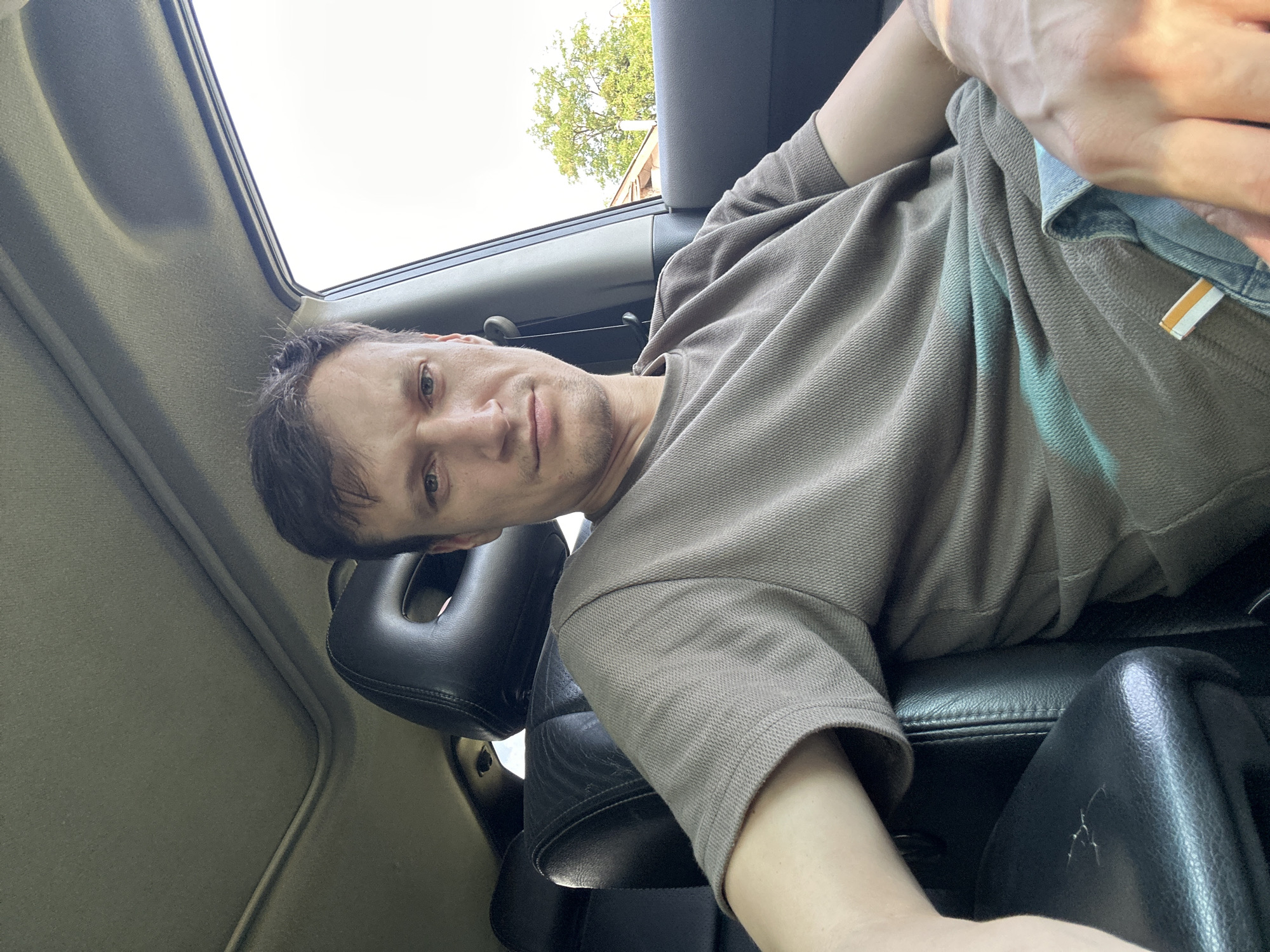 A man is seated in the driver's seat of a car, looking directly at the camera. The interior of the car is visible, with leather seats and a sunroof. Outside the car, trees and a building can be seen. The lighting suggests it is daytime. The man is wearing a brown t-shirt and blue jeans. The location is the city of Makiv, Ukraine.