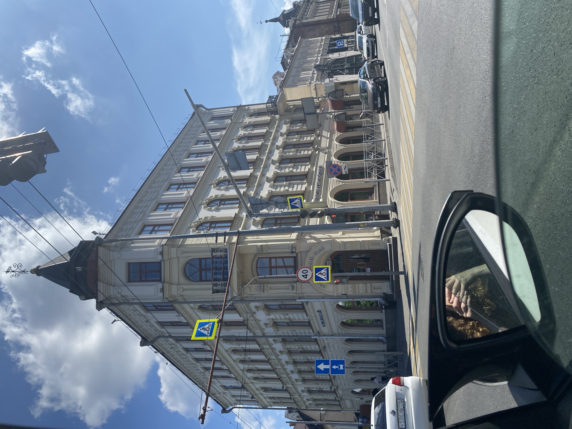 In Kazan, Russia, a perspective shot from inside a car looks out onto a sun-drenched street with a grand, light-colored building dominating the scene. The building, likely an apartment or office building, features multiple floors with arched windows and decorative architectural details. A prominent corner tower with a dark, pointed roof and a weather vane crowns the structure.
Street signs are visible, including a blue and yellow pedestrian crossing sign, a red and white speed limit sign indicating 40, and directional arrows. Traffic lights are also present, with one showing a red light. Several cars are on the road, including a white car in the foreground with a Russian license plate, a dark sedan, and a larger dark SUV. The road surface is asphalt, marked with a pedestrian crossing.
A reflection in the car's side mirror reveals a person with curly hair and hands touching their face, suggesting the photographer. The sky is blue with scattered white clouds, and overhead wires and poles are visible, indicating an urban environment with trolleybus or tram lines. A street name sign on the building appears to read "Кремлёвская" (Kremlyovskaya), confirming the location as Kremlyovskaya Street in Kazan. The overall impression is a sunny day in a historic part of the city.