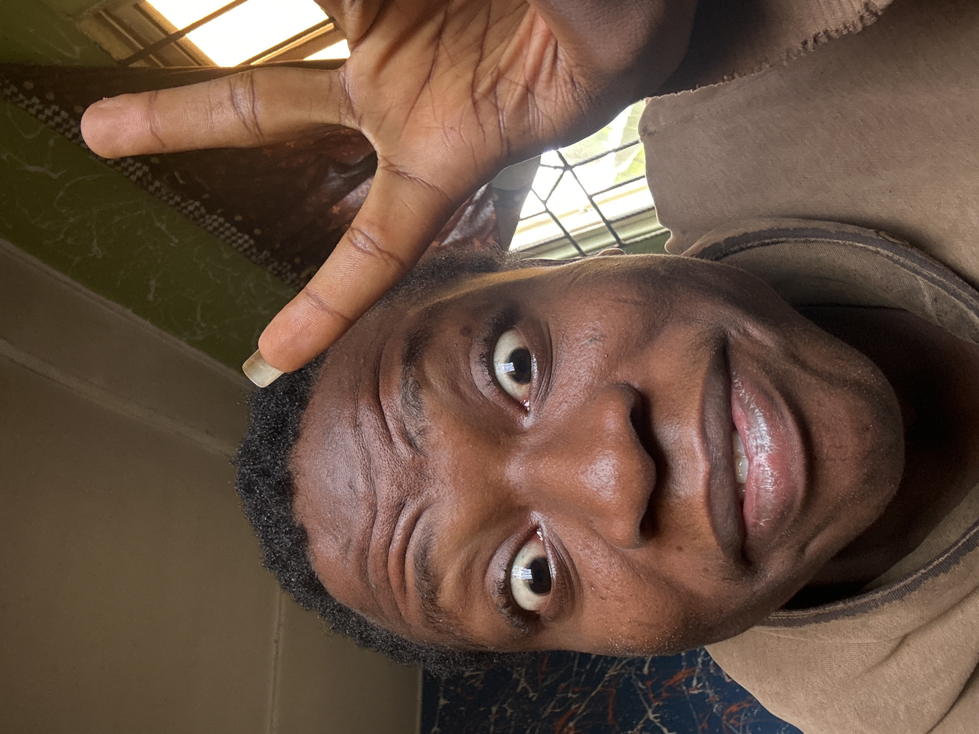 A close-up, tilted portrait shows a dark-skinned person with short, black hair, looking directly at the camera with a slight smile. Their right hand is raised above their head, fingers extended, creating a peace sign gesture. The background includes a window with a lattice-like design, as well as glimpses of patterned surfaces. The person is wearing a brown garment with a raised collar. The location is Unknown, Unknown.