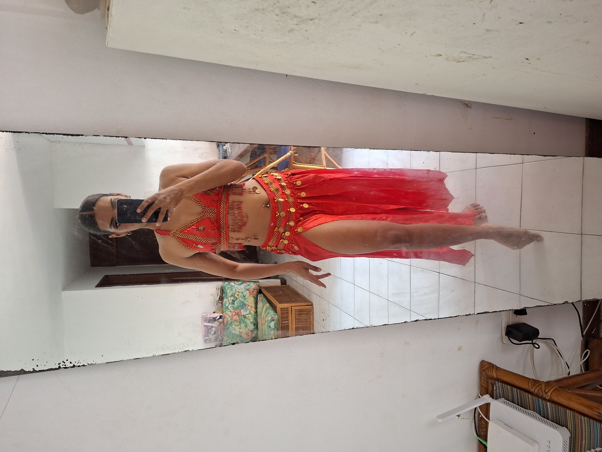 A woman in a red belly dancing costume is standing in front of a full-length mirror in a room. The costume consists of a halter-neck bra adorned with gold beads and chains, and a matching high-slit skirt. A gold coin belt with dangling charms is cinched at her waist. She is holding a black smartphone in her right hand, taking a selfie. Her left arm is extended downwards. The mirror reflects a tiled floor, a wicker bench with a floral cushion, and part of a doorway. In the foreground, to the left of the mirror, is a wooden side table with a white wireless router and cables. To the right of the mirror, a white angled object, possibly part of a shelf or cabinet, is visible. The environment suggests an indoor residential space, potentially in Porlamar, Venezuela, given the tiled flooring and furniture style commonly found in the region.