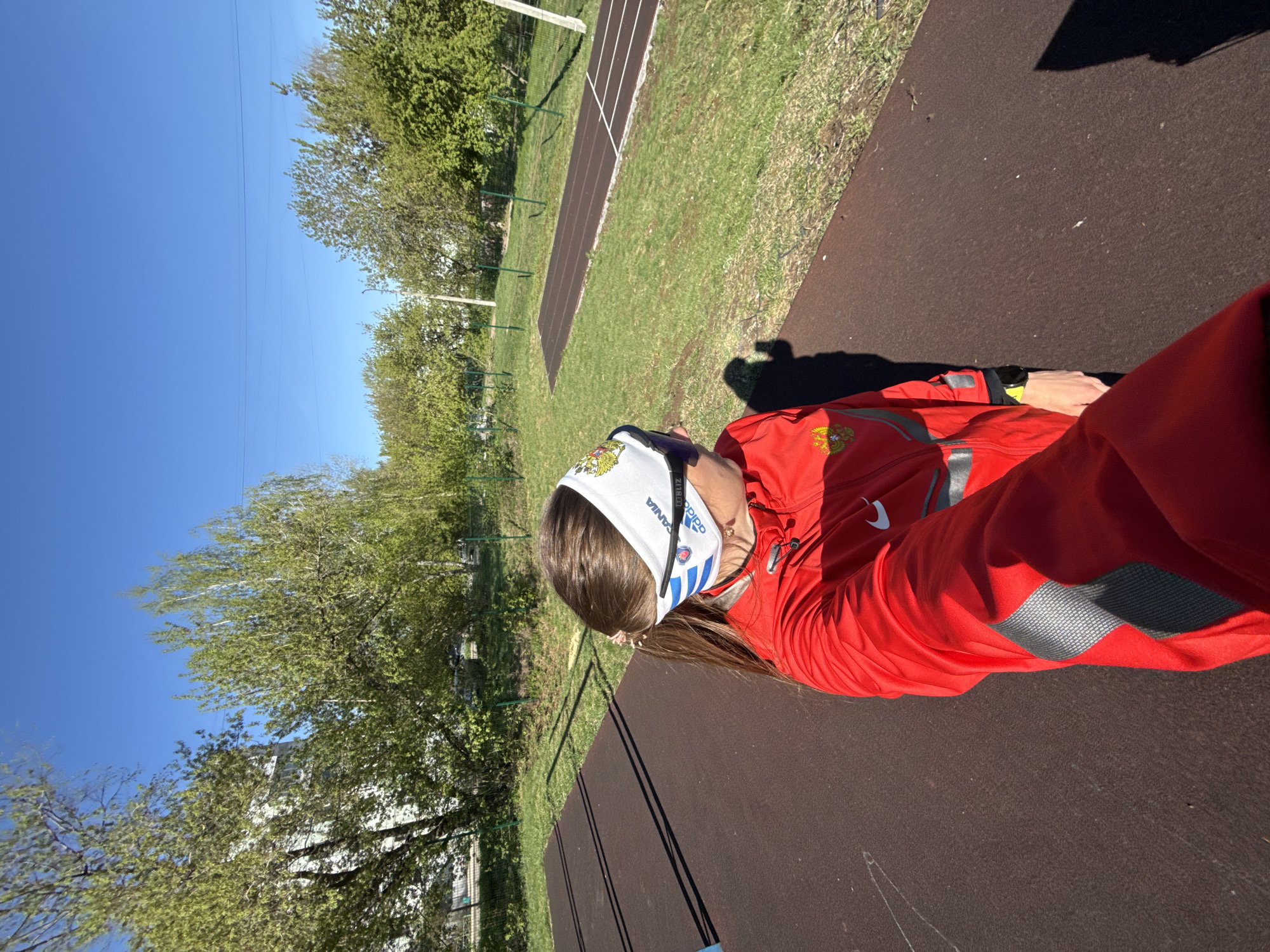 A person wearing a red jumpsuit with reflective accents and a white Adidas headband with blue stripes is standing on a brown, textured surface. The person also wears sunglasses. The location appears to be outdoors, with trees, grass, and a running track visible in the background. The sky is clear and blue. Based on the logo of Russia, the image has likely been taken in the city of Chaikovsky, Russia.