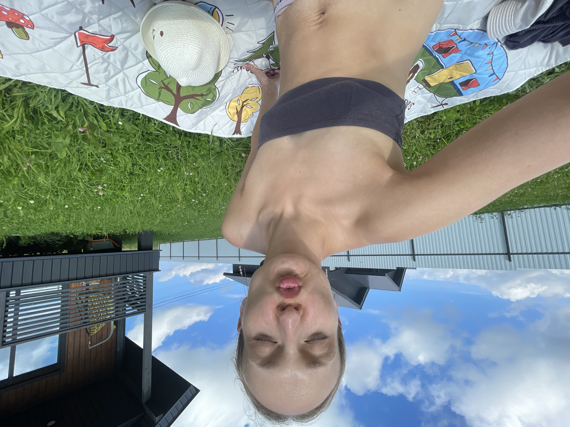 In the foreground, a fair-skinned woman with blonde hair pulled back is taking a selfie outdoors. She is wearing a dark gray strapless top and her eyes are closed, with her lips puckered as if for a kiss. She is sitting on a patterned blanket with cartoonish images of the Earth and various objects. To her right, a white straw hat rests on the blanket. Behind the woman, a green lawn stretches out, dotted with small white and pink flowers. In the mid-ground, a dark metal fence runs horizontally across the image. Beyond the fence, a wooden house with a dark roof and railing is visible. The sky above is a bright blue, filled with fluffy white clouds. The scene appears to be in a rural or suburban setting, possibly in the village of Podshivalovo, Russia, given the presence of a modern house and fence in a grassy, open area.