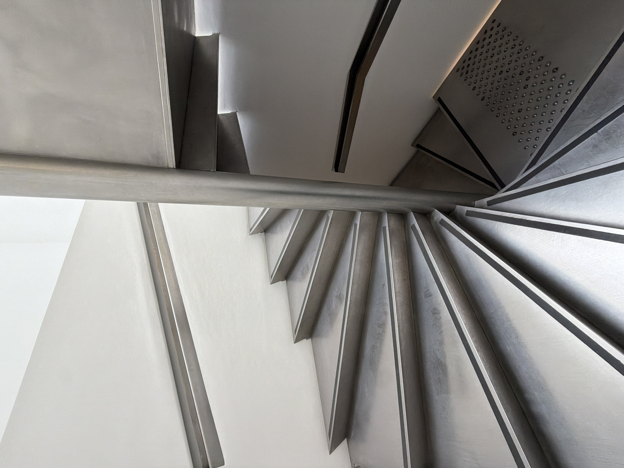 The image depicts a modern, metal spiral staircase viewed from above. The stairs are constructed of a light-colored metal with darker edging, forming a sharp contrast. The staircase wraps around a central column. A metal handrail runs along the side of the stairs. A wall, painted in a light neutral color, is partially visible on the left side of the image. One of the steps is perforated with numerous small holes. The location is undetermined.