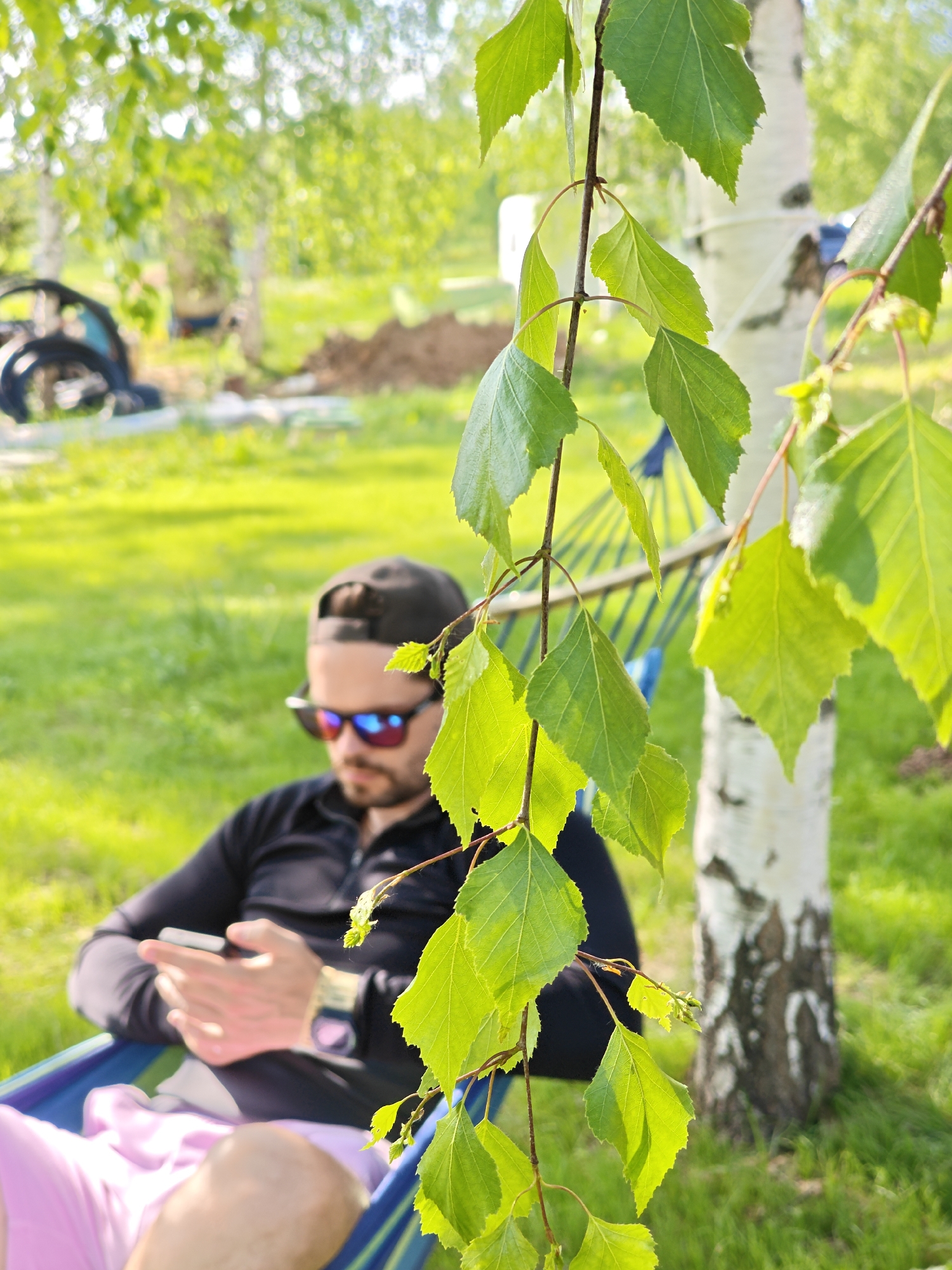 A man wearing a black cap, sunglasses, and a black long-sleeve shirt is sitting in a hammock, looking at his cell phone. He is also wearing a watch, and his legs are clad in pink shorts. The hammock is suspended between two birch trees, with green leaves hanging in the foreground. The scene is set in a grassy area, with trees in the background. The location appears to be Мышкино, Russia, based on the birch trees and general environment. The focus of the image is on the man relaxing in the hammock and using his phone.