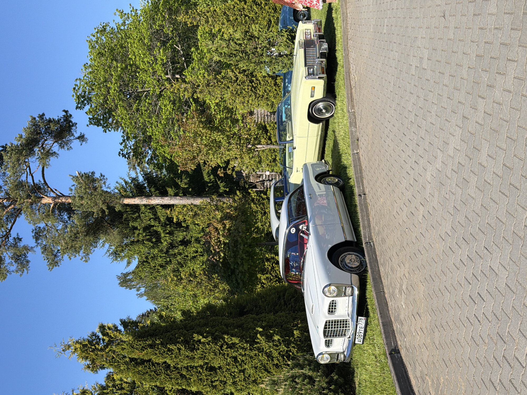 The scene depicts two classic cars parked on a grassy area adjacent to a paved walkway. The car in the foreground is a white convertible with a red interior, prominently displaying a license plate that reads "A089TE778." Behind it, partially obscured by trees, is a light yellow convertible. Lush green trees dominate the background, and a clear blue sky is visible above. The walkway is made of interlocking gray pavers. The location is Repino, Russia, as indicated by the license plate format.