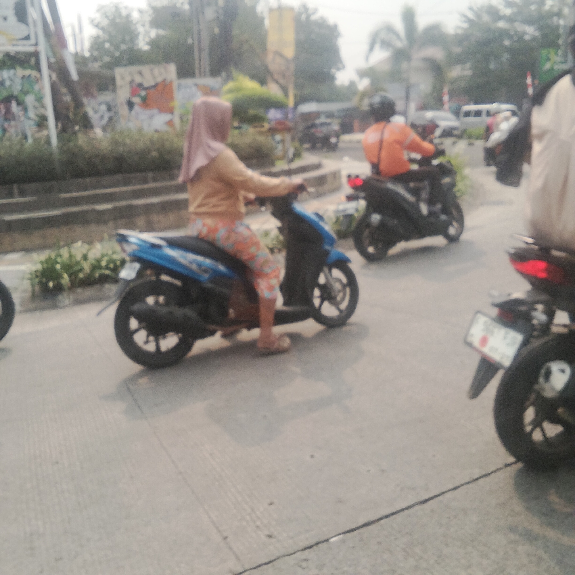 In a street scene in Bogor, Indonesia, a woman wearing a pink hijab and a light brown jacket rides a blue scooter. Her pants are patterned with orange, pink, and blue. She is positioned on the left side of the road. To her right, another scooter is visible, with a rider wearing an orange jacket and a black helmet. Further down the road, a white van and other vehicles are present. The background features greenery, buildings, and overhead power lines, suggesting an urban environment. The image is slightly blurred, indicating motion.