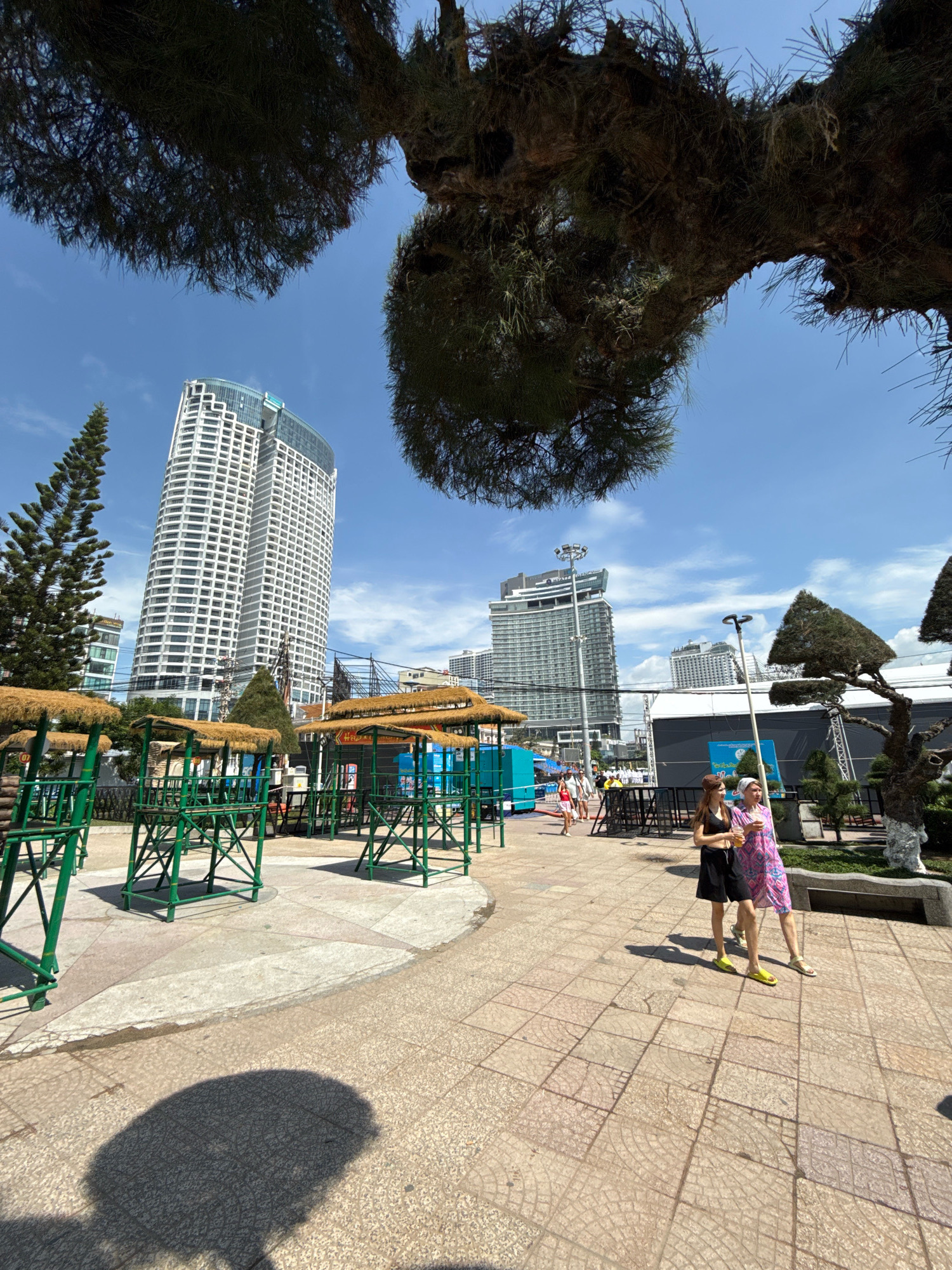 A wide-angle photograph shows a sunny daytime scene with a bright blue sky and scattered clouds. In the foreground, two young women walk side-by-side on a tiled pathway. The woman on the left wears a black tank top and black shorts, while the woman on the right wears a colorful patterned dress and a white headscarf. Both are wearing sandals.
Behind them, an area with green metal structures covered in straw roofs suggests a marketplace or food stalls. Further back, a tall, modern white skyscraper with a curved facade dominates the skyline, its glass-topped roof reflecting the sunlight. Another less prominent skyscraper is visible to its right. The background also shows more buildings, trees, and some scaffolding. The scene appears to be located in Nha Trang, Vietnam, with the distinctive architecture and tropical vegetation suggesting a coastal city environment. Overhead, large tree branches, possibly from a pine tree, frame the top of the image, casting shadows on the pathway.
