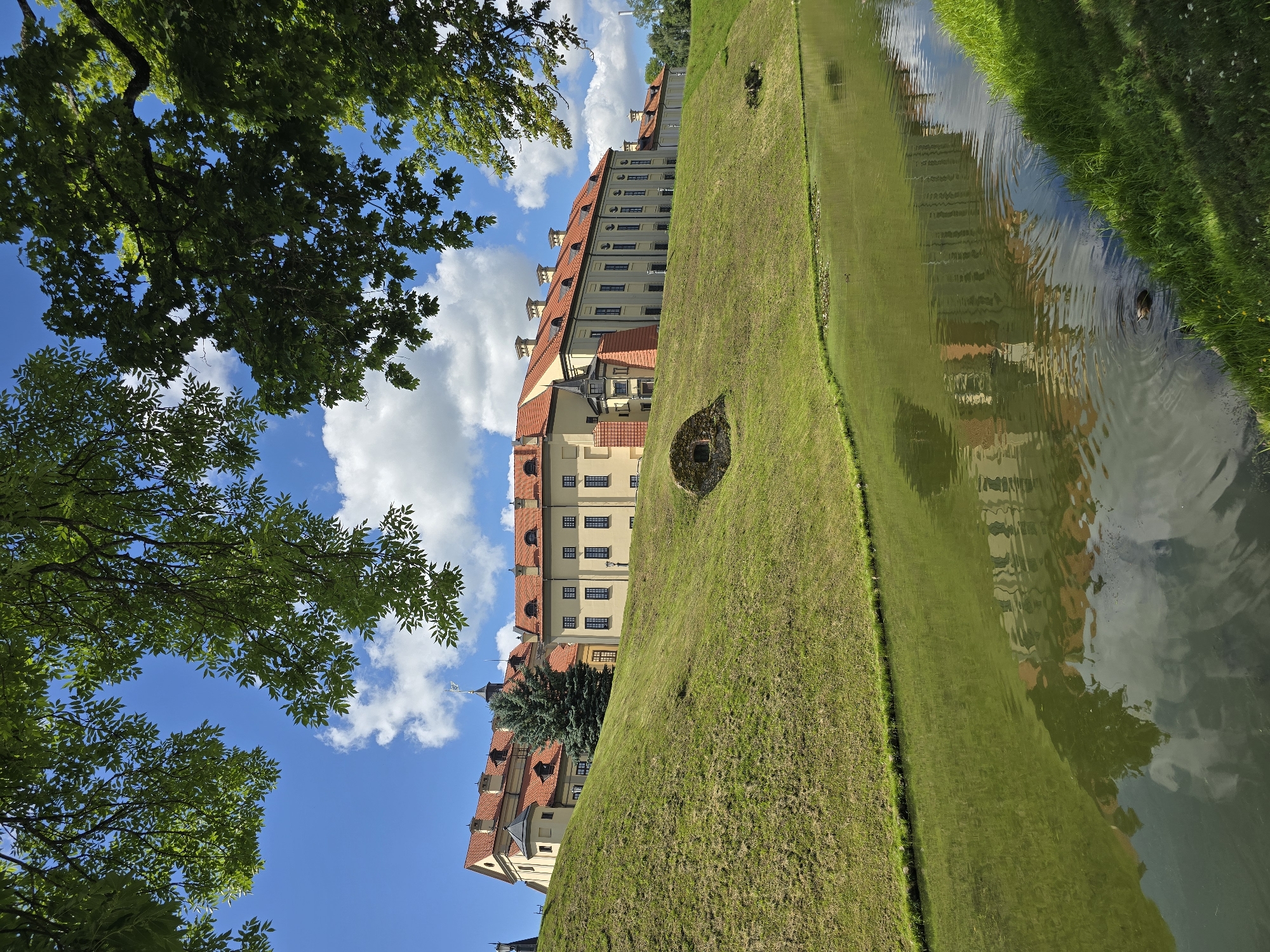 The image shows the Nesvizh Castle complex in Nesvizh, Belarus, on a sunny day with a partly cloudy sky. In the foreground, a calm body of water reflects the castle and the sky. The water is surrounded by lush green grass. A gentle slope of grass leads up to the castle, which is a large, light-colored building with a red tiled roof. The castle has multiple windows and architectural details, including turrets and chimneys. Trees with green leaves frame the top of the image, casting shadows and adding depth to the scene. A small round structure with an arched opening is visible on the grassy slope. There is a single duck swimming in the water.