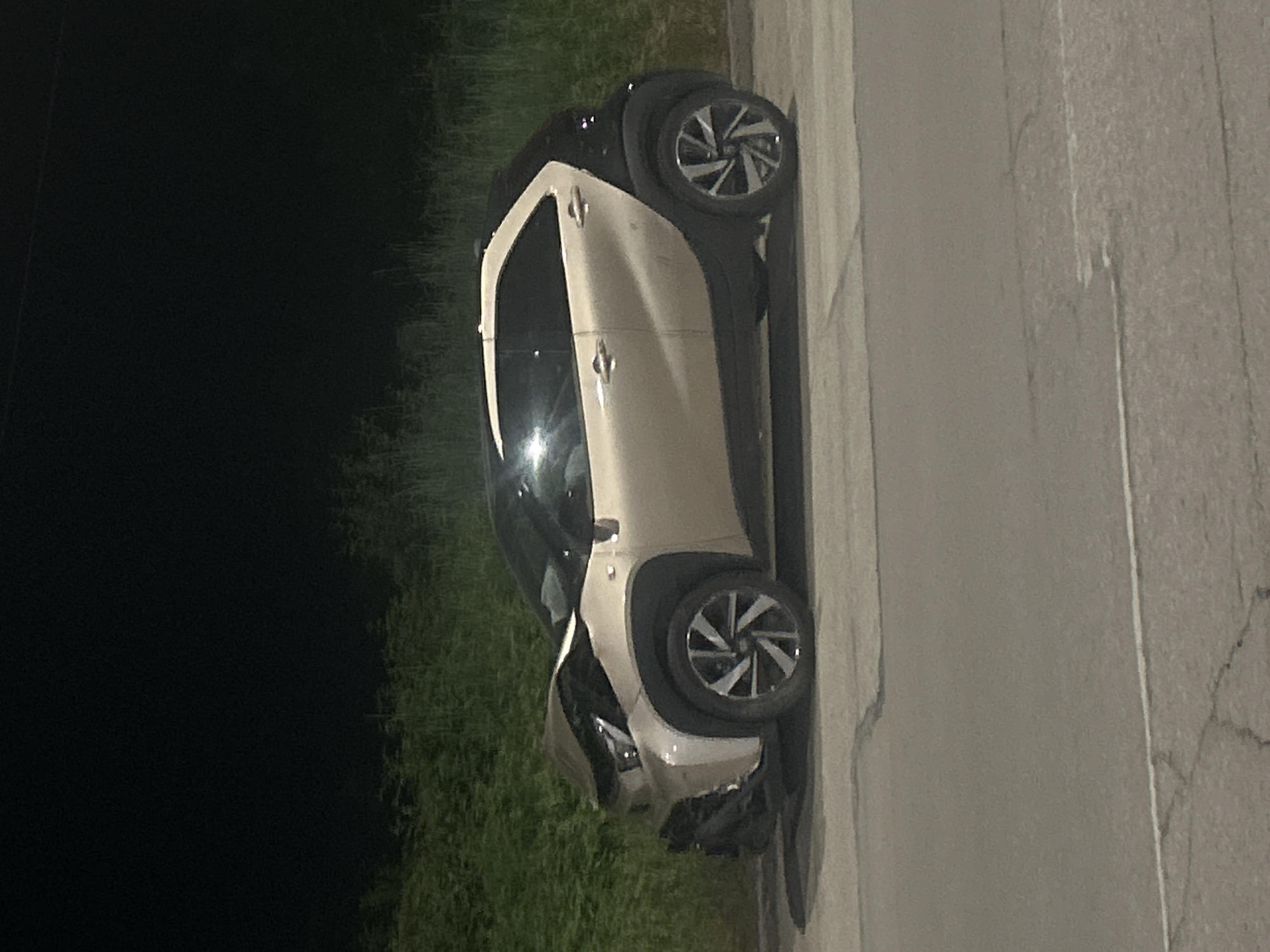 A two-toned compact car, primarily beige with black accents, is parked partially on a cracked, asphalt road and partially on the grassy shoulder. The front end of the vehicle is significantly damaged. The scene appears to be at night with dark surroundings except for the road and the car illuminated by an unseen light source. The location is within an urban area, evidenced by the presence of the paved road and nearby vegetation, but the specific location within the city of Pleven, Bulgaria, is indeterminable from the visual information provided.