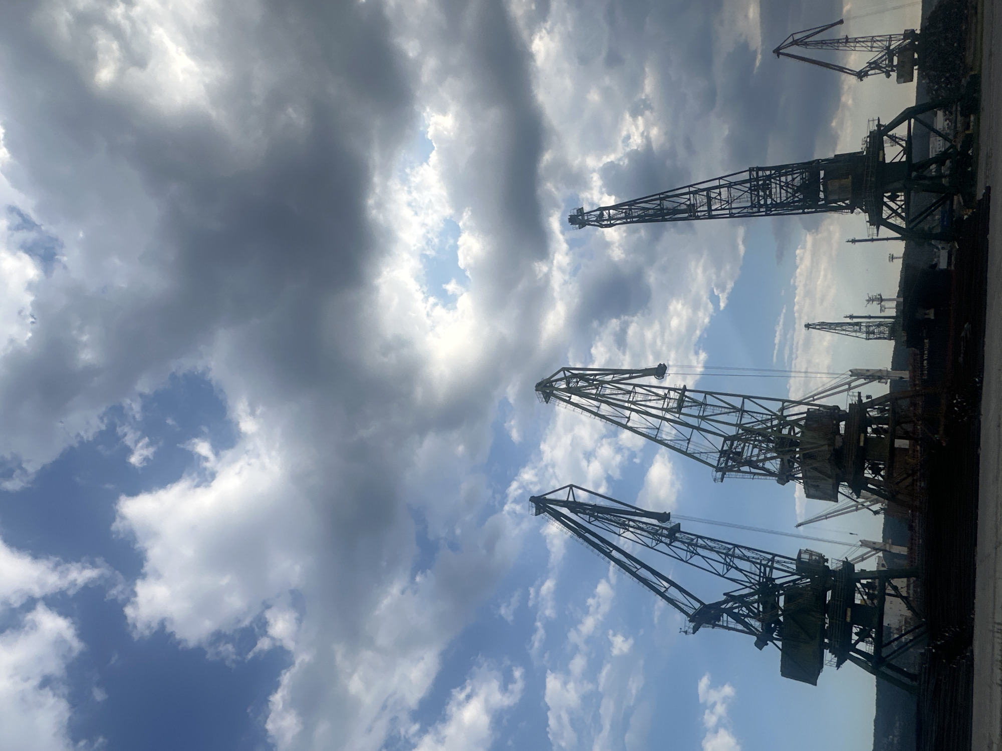 This is a low-angle, full-shot photograph of a harbor scene with multiple large industrial cranes dominating the frame. The sky above is a mix of dramatic, heavy grey clouds and patches of bright blue sky with scattered white clouds. The cranes, some appearing weathered and industrial green, are silhouetted against the sky. The closest crane on the left is a massive structure with a prominent boom. To its right, another large crane is visible, also with a substantial boom and cab. In the background, several other cranes of varying sizes are scattered across the landscape, some with red accents. The base of the cranes appears to be situated on a dock or a large ship, with a dark, textured foreground suggesting a pile of materials or a harbor floor. The overall impression is one of industrial activity under a dynamic, cloudy sky.