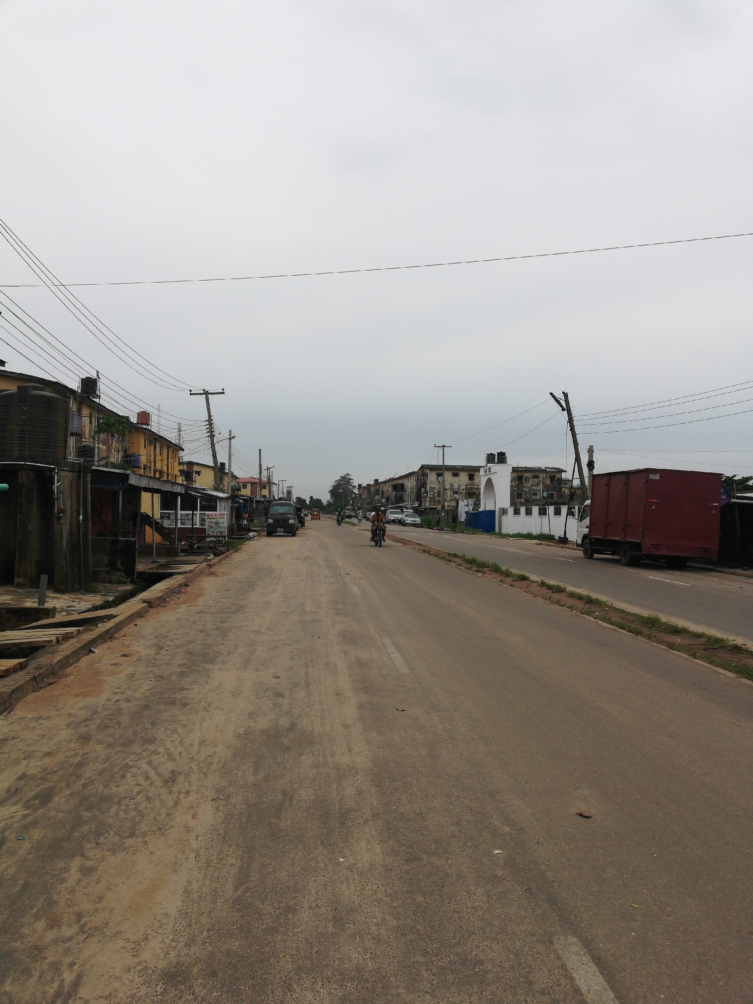 An eye-level, long shot photograph depicts a partially unpaved street in Lagos, Nigeria, under an overcast sky. The street, divided by a painted white line, stretches into the distance, with a mix of vehicles and pedestrians visible. The left side of the street features older, weathered buildings, some with water tanks on the roofs and open-front businesses. A dirt and debris-filled gutter separates the buildings from the road. Power lines run overhead. The right side of the street shows newer buildings, including a white building with an arched entrance. A large red shipping container on the back of a truck is parked along this side. Several people are riding motorcycles and walking along the street.