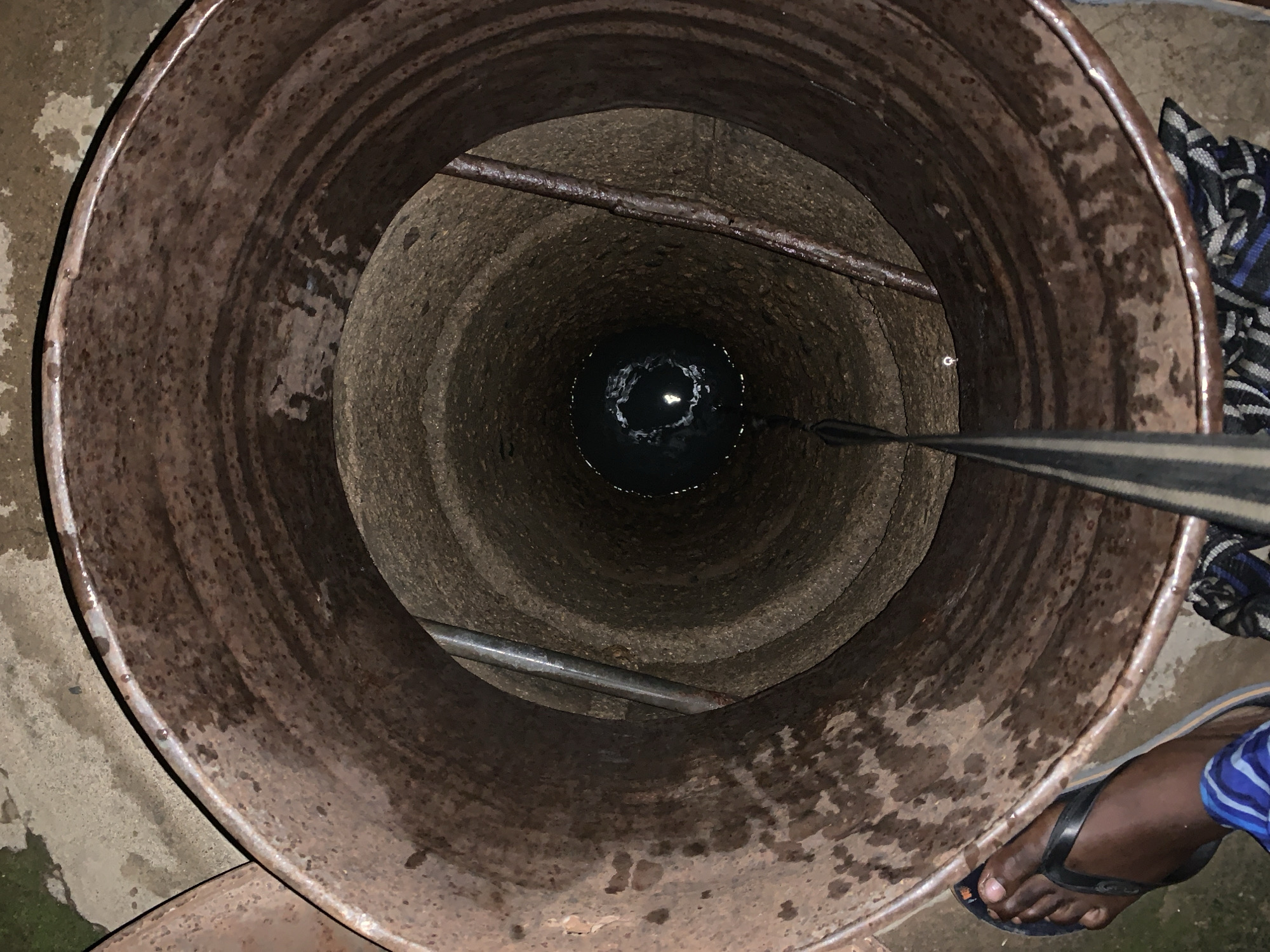 The image provides a top-down view into a deep well located in Rafin Par, Nigeria. The well's circular opening is surrounded by a large, rusty brown metal casing or drum. The inner walls of the well shaft are dark, textured, and appear to be constructed from concrete or stone rings, showing signs of dampness and wear.
Two horizontal metal bars are visible across the well's interior at different depths, likely serving as structural support. At the very bottom of the shaft, a small pool of water reflects a bright light source from above. A wide, dark-colored woven strap or rope descends into the well from the upper right side, indicating its use for drawing water.
In the lower right portion of the frame, a dark-skinned foot wearing a black flip-flop sandal is visible near the edge of the well, suggesting the presence of a person engaged in water collection. A piece of patterned fabric, black with blue and white accents, is partially draped near the well opening on the right.
The scene depicts a practical activity of accessing water from a well, a common daily task in many communities. The well's interior is dark, but the reflection on the water at the bottom indicates the presence of an overhead light source, possibly natural daylight or an artificial light. No visible text is present in the image.
