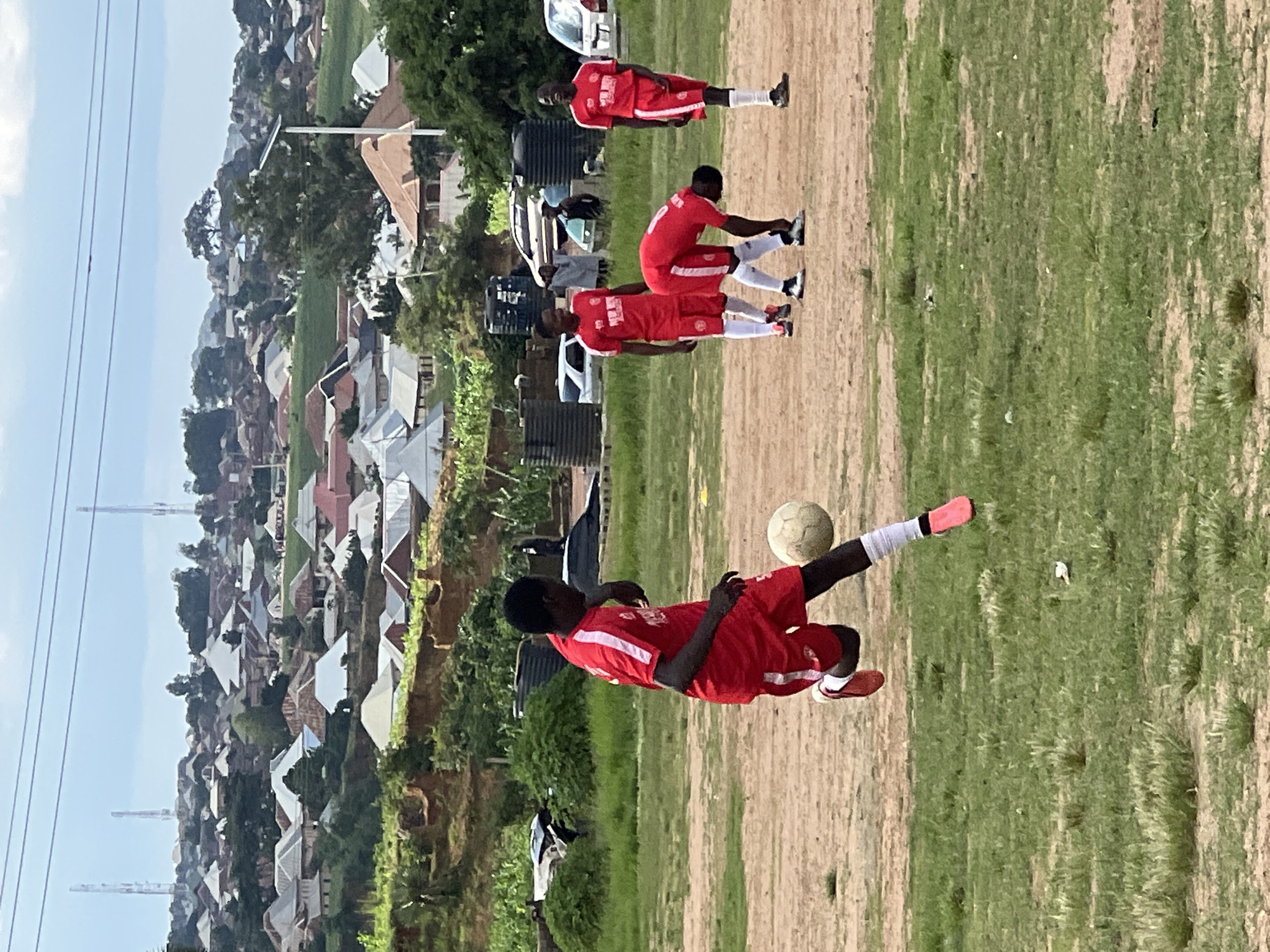 The image depicts a group of young men in red soccer uniforms on a dirt field in Jos, Nigeria. In the foreground, a player dribbles a soccer ball, his body angled as he prepares to kick. Several other players in similar uniforms are scattered across the field, one bent over, another standing, and one in the mid-ground looking towards the foreground player. The background reveals a densely populated hillside with numerous houses, varying in color and architectural style, some with corrugated iron roofs. Communication towers and a solar-powered street light are visible in the distance, suggesting an urban or semi-urban environment. The terrain is uneven, with patches of dry grass and exposed dirt. The sky is overcast, indicating a cloudy day.