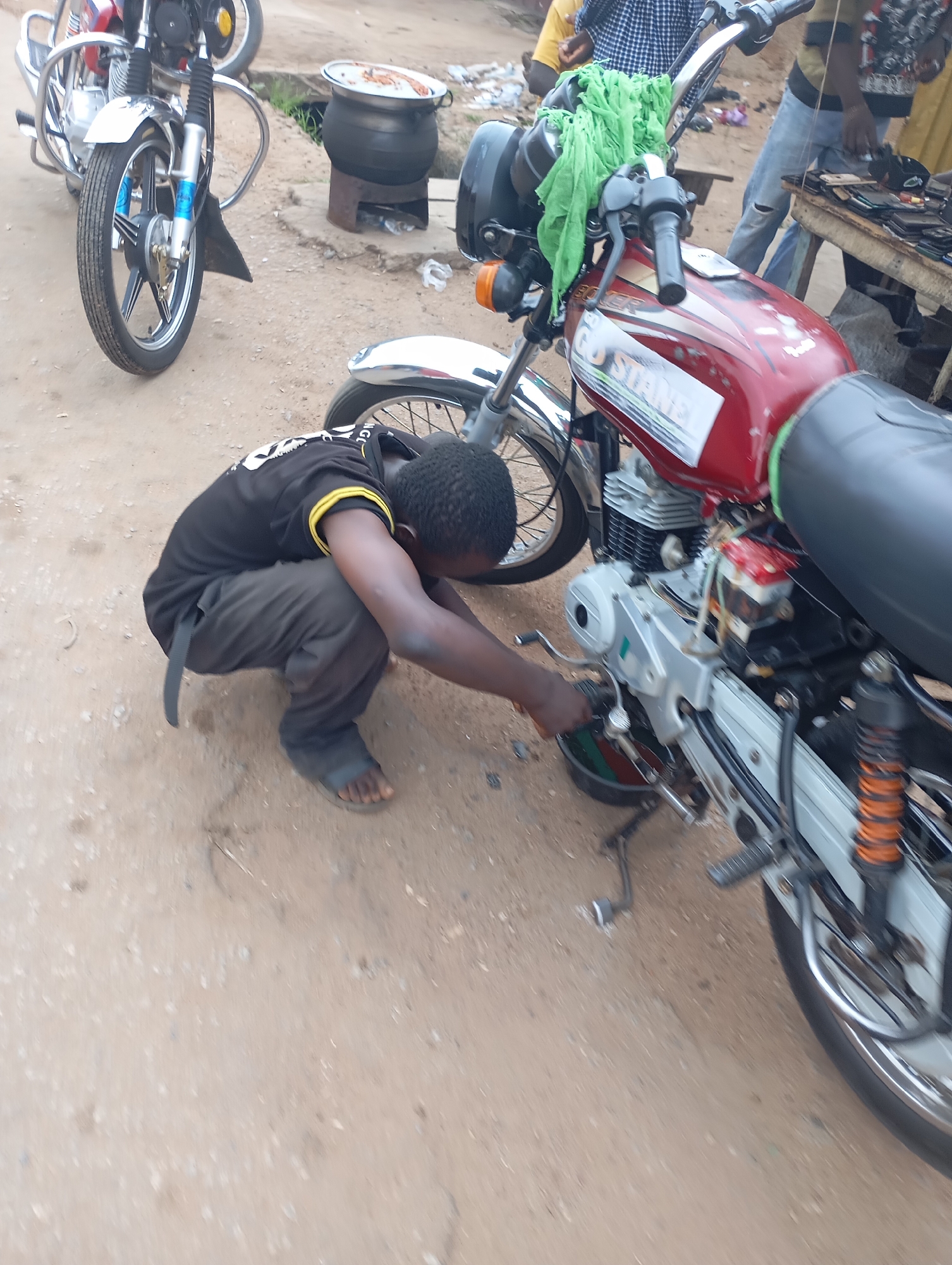 In Shendam, Nigeria, a young boy in a black t-shirt and dark pants crouches to work on a red motorcycle. The motorcycle is parked on a dirt road, with other motorcycles and a large pot on a stove visible in the background. The boy appears to be focused on the engine of the motorcycle, with his hands actively engaged in the repair. The scene suggests a typical day in a Nigerian city, with a blend of commerce and daily life unfolding on the street.