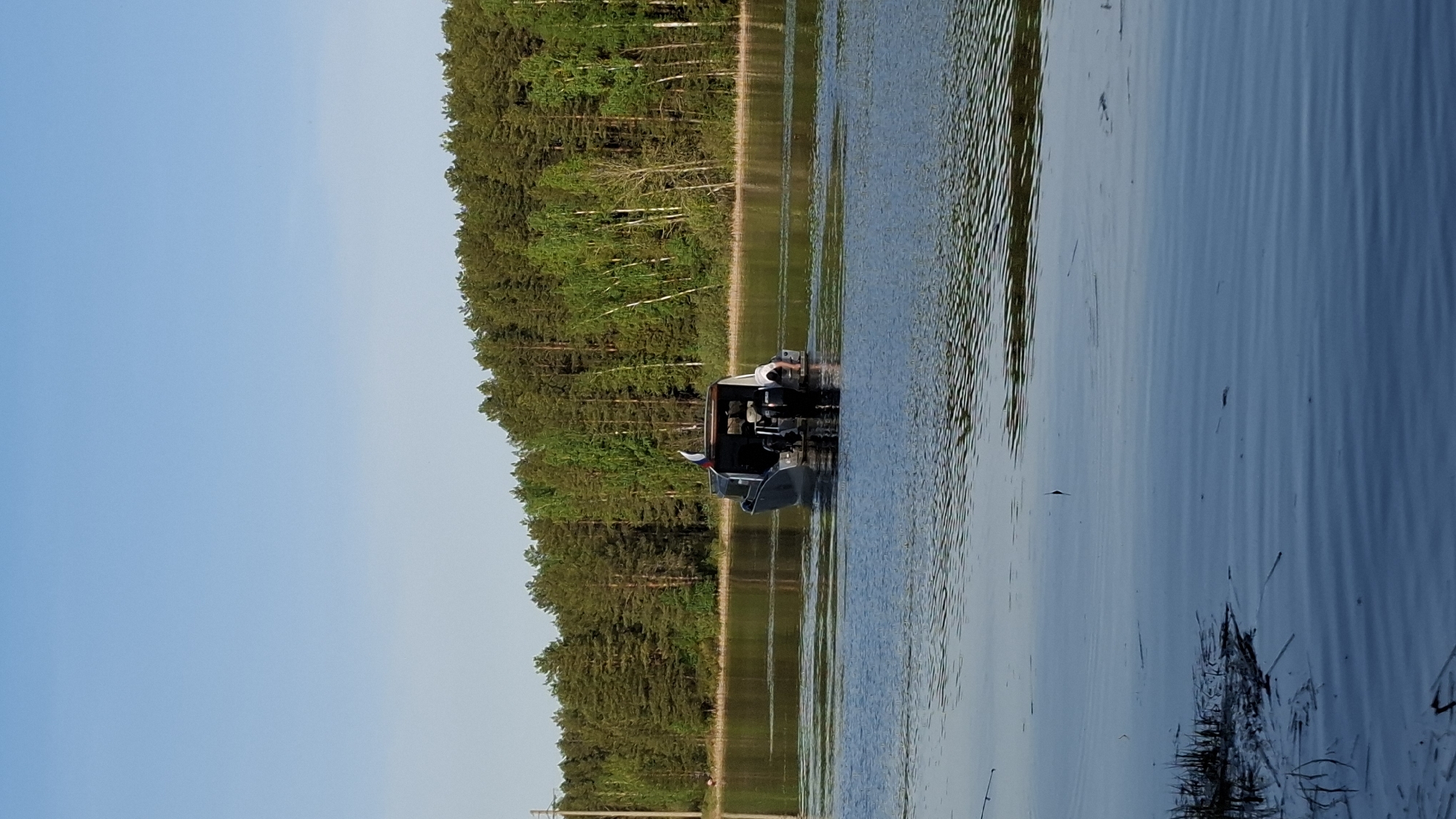 The image shows a motorboat with a person working on the motor. The motorboat is in the water, and there is a forest along the shore in the background. The water appears to be a lake or a river. The sky is blue. The location is near the city of Chaikovsky, Russia, based on the presence of a Russian flag on the boat and the typical landscape of the area.