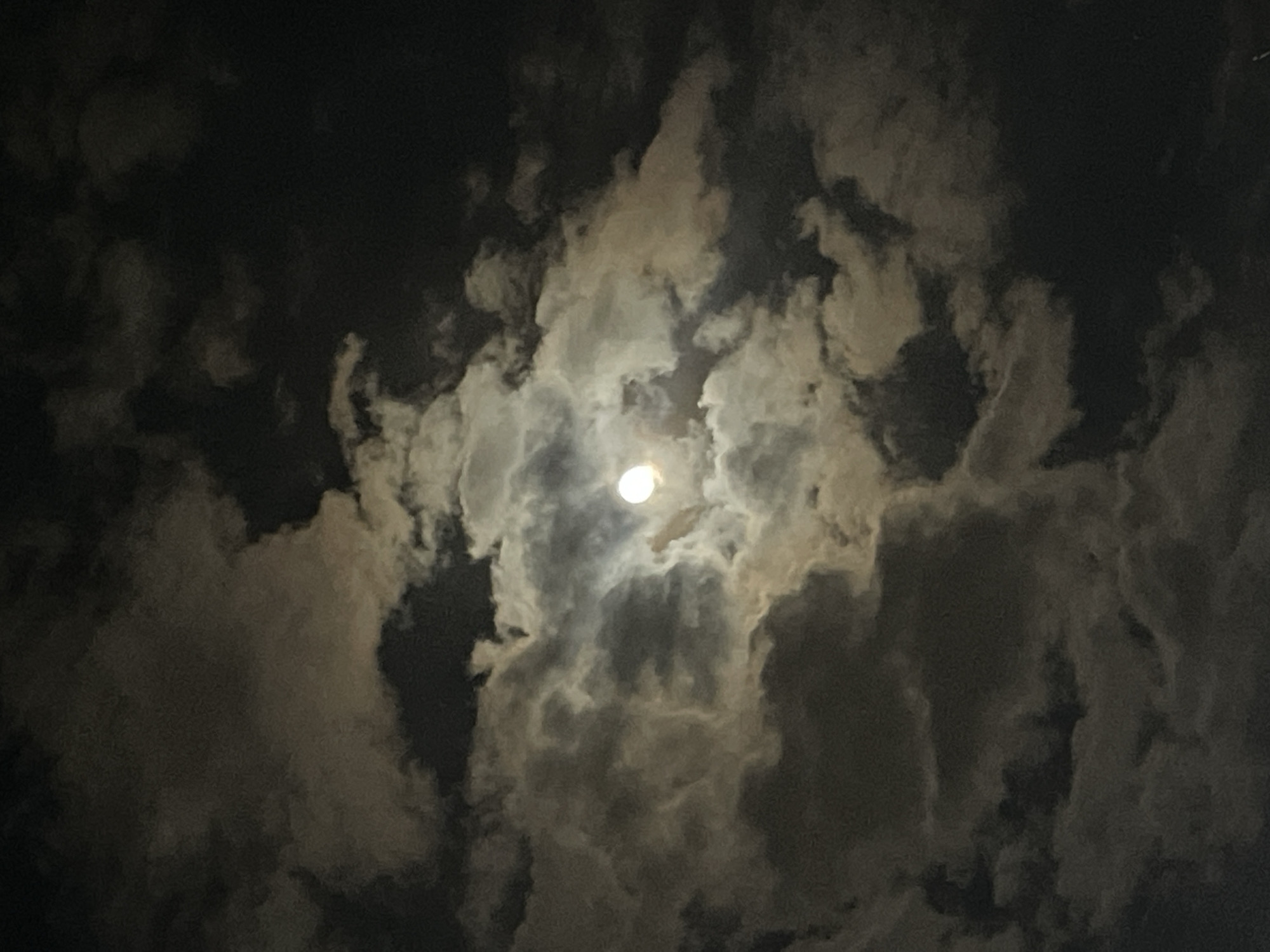 The image depicts a night sky filled with clouds partially obscuring the moon. The moon is positioned towards the center of the frame and shines brightly. The clouds are varying shades of gray, indicating different thicknesses and densities. The environment appears dark, suggesting a nighttime setting. There is no information to determine it is taken in Yerevan, Armenia.