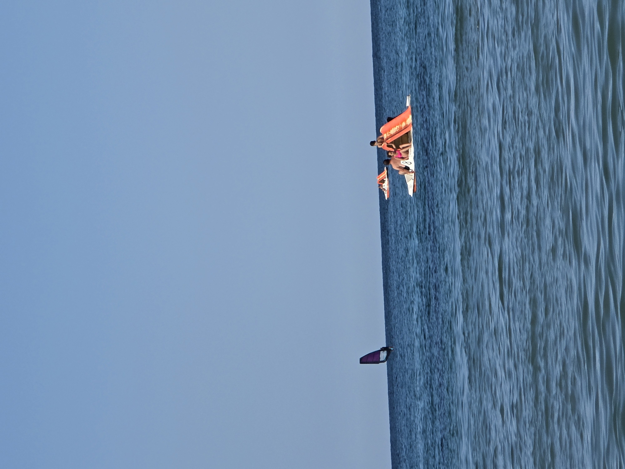 The image depicts a clear, sunny day at a coastal location, likely Ahtopol, Bulgaria, with a vast expanse of blue sky meeting a tranquil blue sea. In the distance, a lone windsurfer navigates the water, their purple sail a striking contrast against the horizon. Closer to the foreground, two paddle boats are visible. One paddle boat carries a group of people enjoying a leisurely outing. Another paddle boat is occupied by a family; a man and a child are seated on the edge, while a woman and a young girl are on the slide, with the girl in a pink swimsuit. The water is calm with gentle ripples, suggesting a peaceful day at the beach.