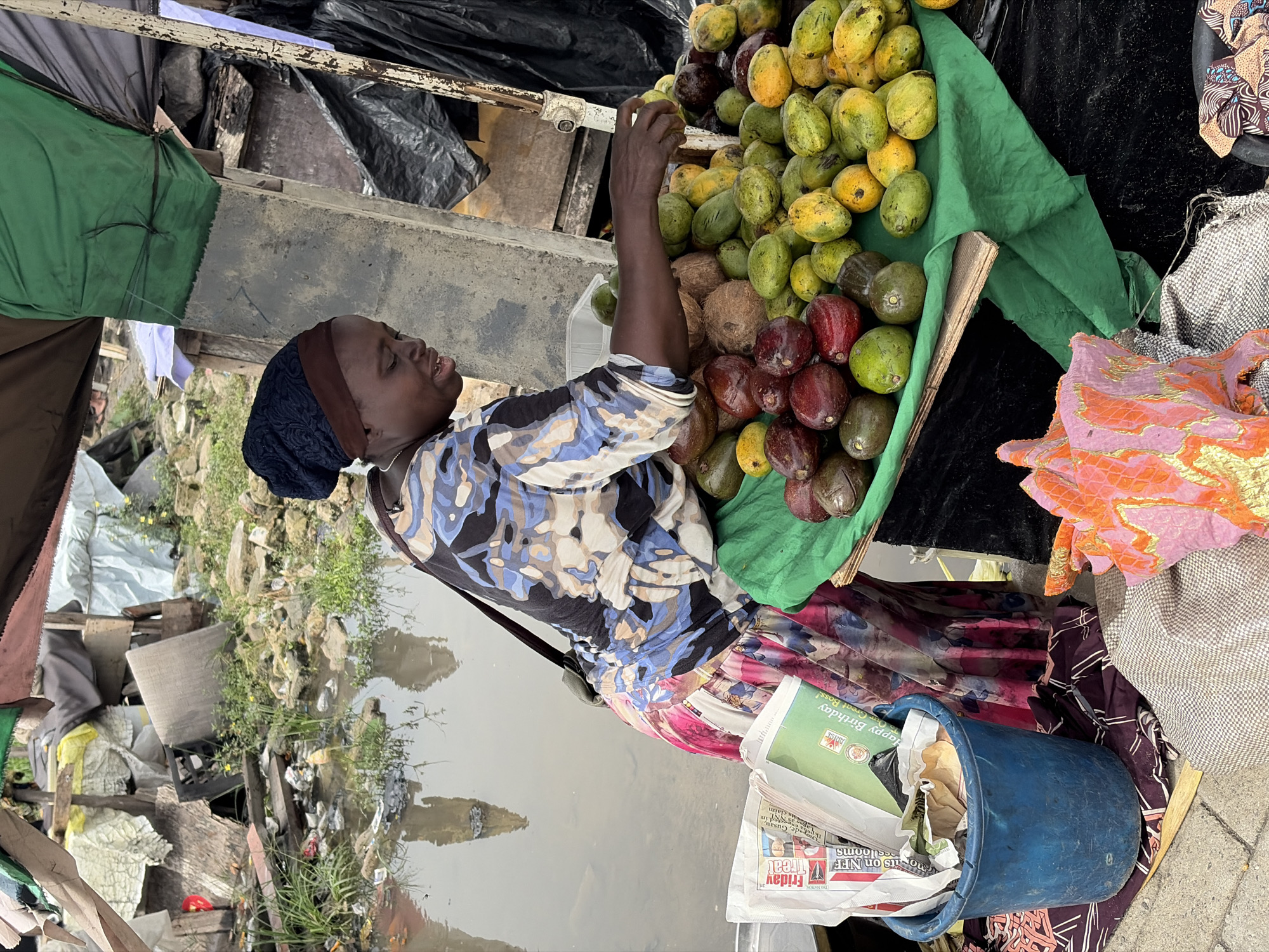 A woman wearing a blue and white patterned shirt, a colorful skirt, and a dark head covering is standing at a fruit stand in what appears to be a market in Igboefon, Nigeria. She is reaching into a pile of fruit, including mangoes, avocados, and coconuts, which are displayed on a green cloth. To the right, there is a blue bucket filled with newspapers. Behind the stand, there is a body of water with visible trash and debris. The overall scene depicts a typical day at a market in a developing urban area.