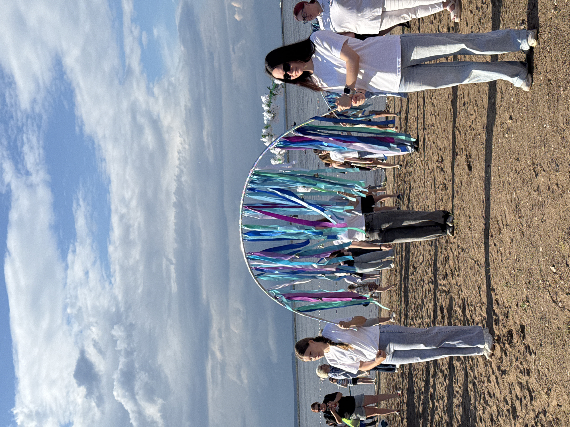 The image depicts an outdoor gathering on a sandy area near a body of water, likely a river or lake, with a cloudy sky overhead. The location is identified as Chaikovsky, Russia. In the foreground, two young women are holding an arched structure decorated with various colored ribbons in shades of blue, purple, and teal, with small silver bells attached. The woman on the left is wearing a white t-shirt and light blue jeans, and the woman on the right is wearing a white t-shirt, sunglasses, and light blue jeans. Behind them, a crowd of people is visible, some standing and some walking. The overall atmosphere appears to be a public event or celebration taking place outdoors, possibly on a beach or riverbank.