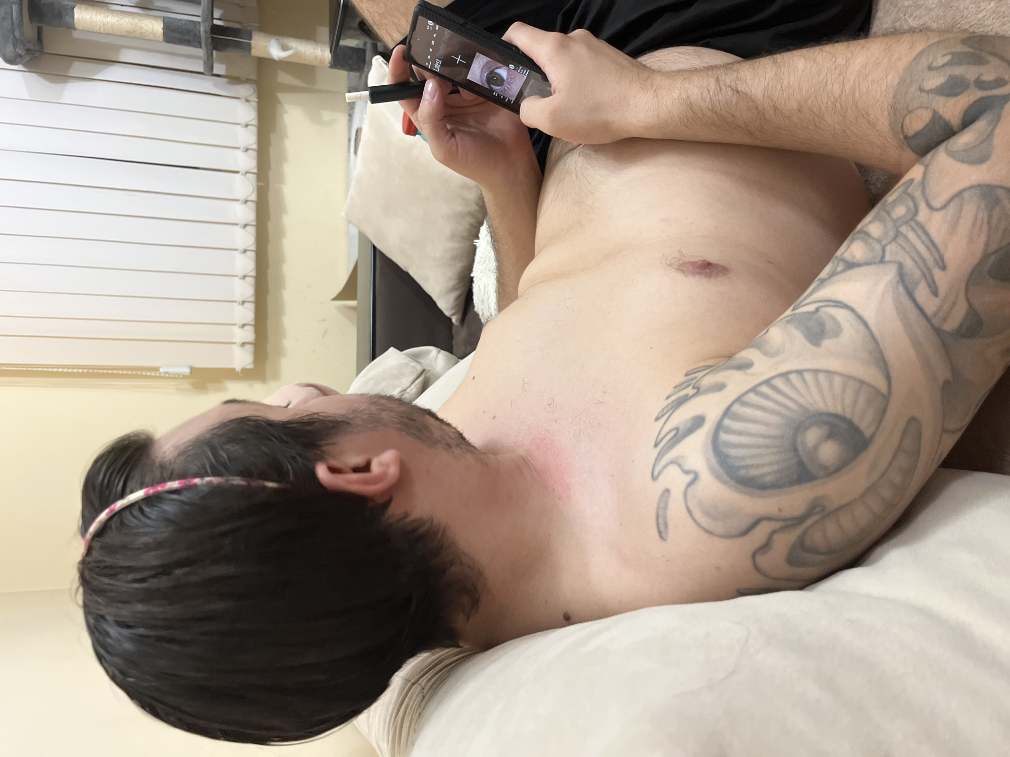A shirtless person is lying on a couch, holding a smartphone in their right hand. The smartphone displays an image of an eye. A tobacco heating device is held in the person's left hand. The person has tattoos on their arm and chest. The environment appears to be a living room, with a cat tree and vertical blinds visible in the background. The location is Plovdiv, Bulgaria.
