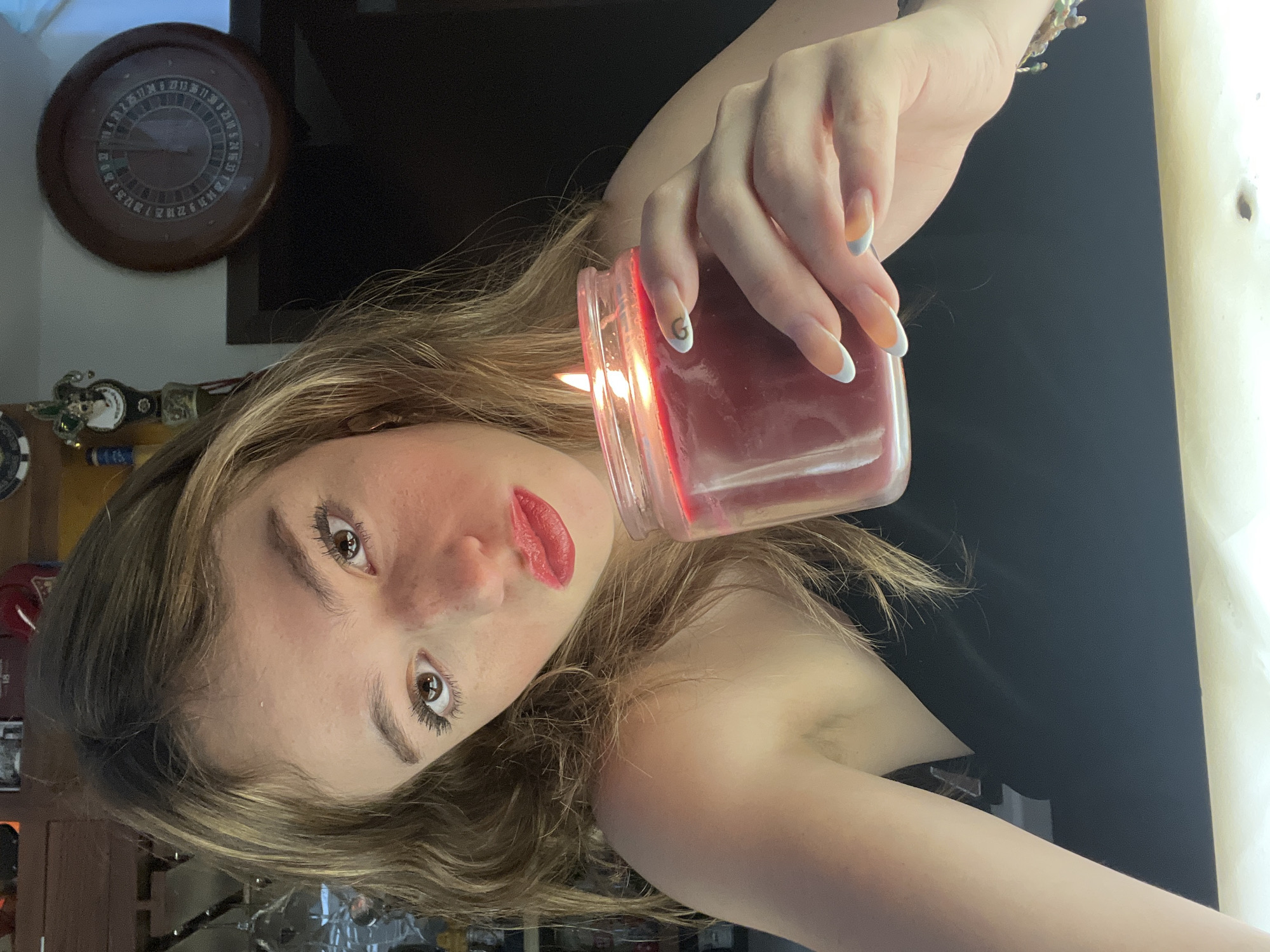 A young woman is captured in a close-up, diagonal shot, holding a lit candle in a red-tinted glass jar. She has long, light brown hair, fair skin, and is looking directly at the camera with dark, expressive eyes. Her lips are adorned with red lipstick, and she wears dark mascara. She appears to be wearing a dark, possibly strapless or off-shoulder top.
Her right hand, positioned prominently, holds the candle jar, with a clear flame visible inside. Her nails feature a French manicure style with white tips; one fingernail (index finger) has a small letter "G" visible, and some nails have a light blue or grey base under the white tip. A beaded bracelet is on her right wrist.
The background features a dark, flat surface, likely a television or screen, behind which a round, wooden object resembling a roulette wheel is mounted on a light-colored wall. The roulette wheel displays various numbers, including 1, 3, 5, 6, 7, 8, 9, 10, 11, 12, 13, 14, 16, 17, 18, 20, 22, 23, 24, 25, 26, 27, 28, 29, 30, 31, 33, 34, 35, and 36. Below it, several small, indistinct items are visible, possibly decorative or gaming-related. To the far right, a light cream-colored surface is visible, potentially a wall or curtain.
The overall mood is contemplative or posed, possibly for an artistic portrait, with the candle providing a soft, warm light source. The image provides no specific visual cues to the provided location of San Bernardino Tlaxcalancingo, Mexico.