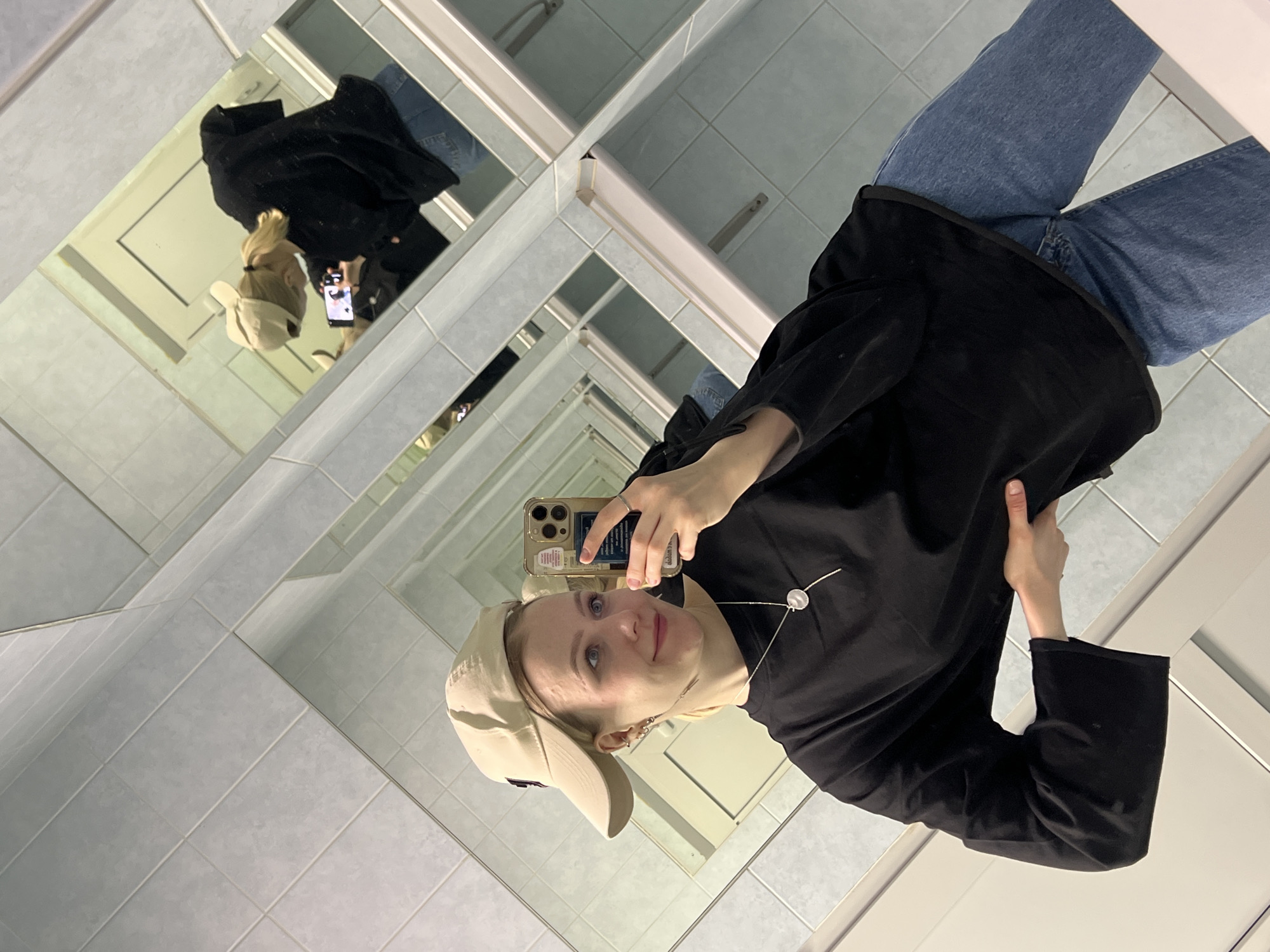 A young woman is captured in a mirror selfie within an indoor setting in Khimki, Russia. She appears to be in her late teens or early twenties, with light-colored eyes and blonde or light brown hair pulled back, mostly concealed by a light-colored (possibly beige) baseball cap. She is wearing a black long-sleeved top or light jacket and blue jeans. Accessories include a silver necklace with a circular pendant and a ring on her left hand.
The woman is holding a smartphone, encased in a gold or beige cover, with her right hand to take the photo, while her left hand is subtly positioned near her hip, possibly adjusting her top. She has a slight, soft smile and her gaze is directed towards her phone's camera in the reflection.
The background consists of light blue-grey tiled walls and floor, typical of a public restroom or changing room. Large mirrors are arranged to create an illusion of infinite reflections, showing the woman and the tiled room receding into the distance multiple times. This mirror effect is a prominent visual element, adding depth and a unique perspective to the image. The lighting is artificial and consistent, indicating an indoor environment. No decipherable text is visible on her phone screen or cap.
