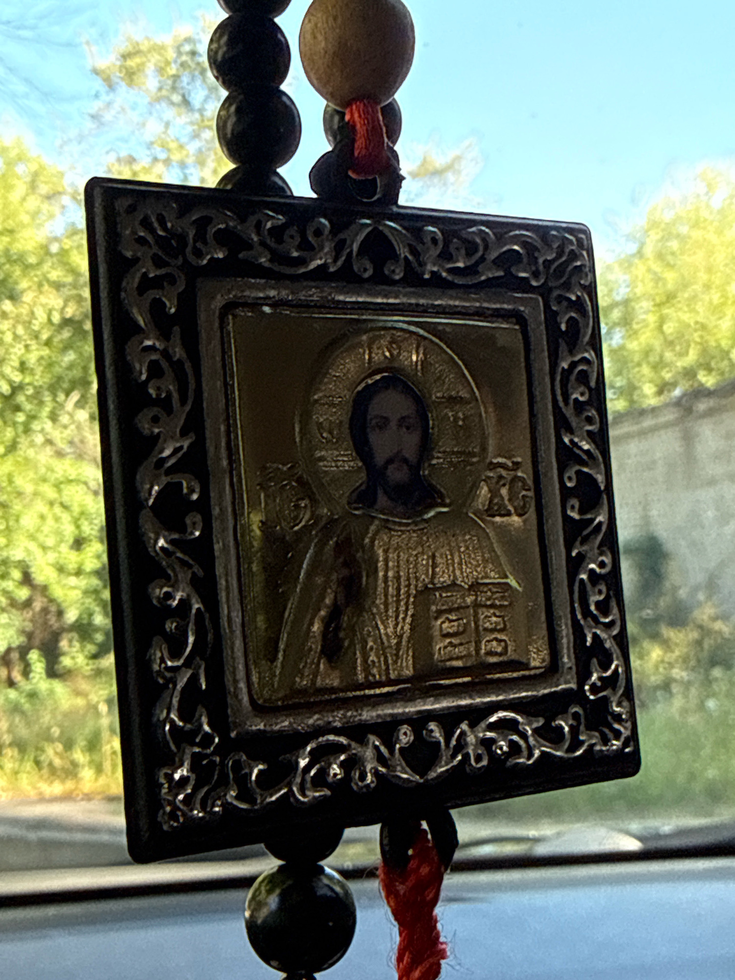 An icon of Jesus Christ hangs in front of a blurred background of trees and sky. The icon is square and framed by a decorative black border with silver accents. Below the icon, there are dark beads and a red cord. The icon depicts Jesus with a beard and halo, wearing a robe. The background suggests an outdoor setting, possibly a car interior, given the way the light is filtering through.