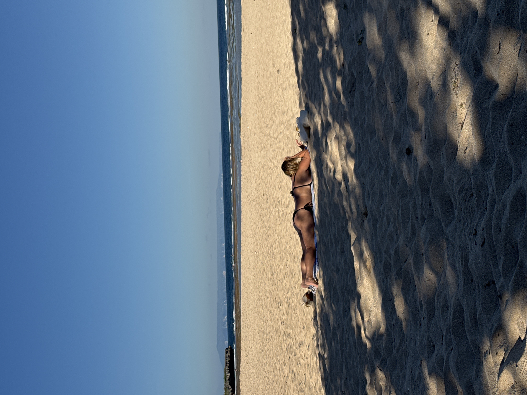 The image shows a woman lying on the beach at Pantai Tegal Wangi in Kuta Selatan, Bali, Indonesia. The woman is wearing a dark-colored bikini and is lying on a light-colored towel on the sand. A pair of sandals sits near her feet. The sand is light in color and textured. The ocean is visible in the background, with the sky a clear blue. Strong shadows suggest the presence of trees.