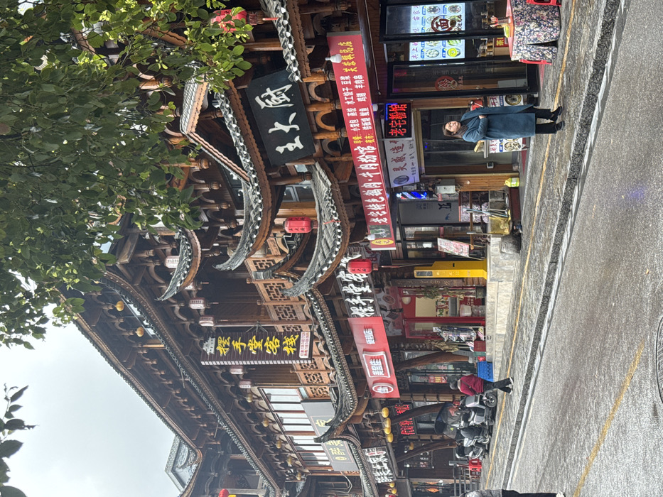 The image depicts a street scene in Furong, China, under an overcast sky. A wet paved street surface occupies the foreground, bordered by a concrete curb and a sidewalk. A tree with green foliage partially extends into the upper right portion of the frame.
The background features a row of multi-story buildings constructed in traditional Chinese architectural style, characterized by dark wooden framework, ornate railings, and multiple tiers of dark-tiled roofs with upturned eaves. Numerous Chinese character signs adorn the facades, including a large yellow vertical sign reading "摆手堂客栈" (Bai Shou Tang Inn) and a black horizontal sign above an entrance displaying "王大厨" (Chef Wang). Other storefronts at street level include a "美滋生便利店" (Mei Zi Sheng Convenience Store) and a "老宅特色牛肉粉面" (Old House Special Beef Noodles) shop, with a barbershop also visible. Red cylindrical lanterns and smaller yellow spherical lanterns are affixed to the buildings.
On the sidewalk in the mid-ground, a woman in a long, dark blue coat stands with her arms crossed, facing right. Further left, a person wearing a red jacket walks away from the viewer. Three scooters or motorcycles are parked parallel to the curb on the left. A bright yellow freestanding machine, resembling a payment terminal, is positioned on the sidewalk in front of a shop. To the far right, a small, round table covered with a floral fabric tablecloth is set up on the sidewalk next to a storefront.