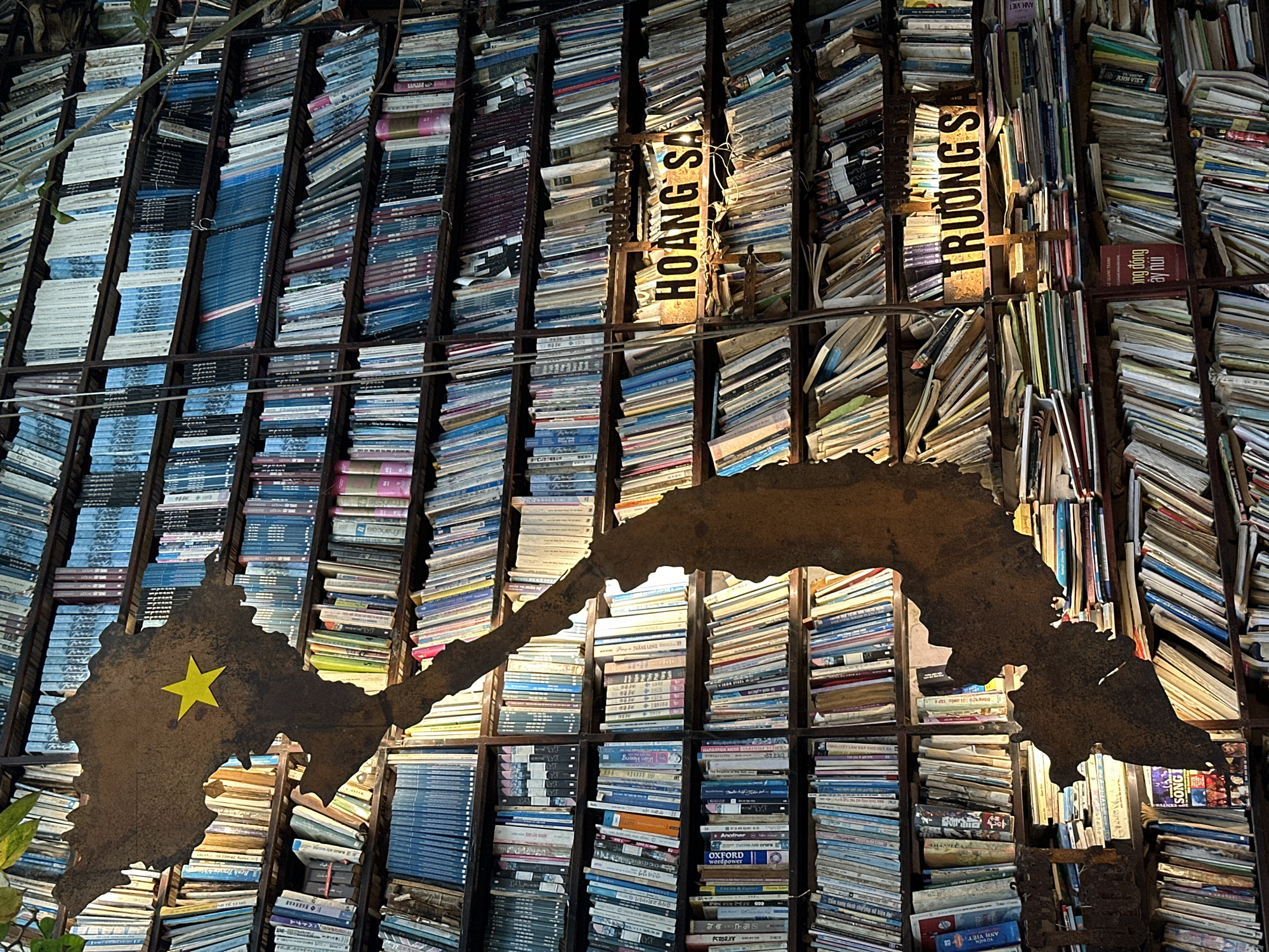 The image shows a book display in Thủ Đức, Vietnam. A rusty metal frame divides the scene into vertical columns, each filled with stacked books, predominantly blue and white. The metal frame supports illuminated signs reading "HOANG SA" and "TRƯỜNG S." A large rusty metal cutout of the map of Vietnam is visible, adorned with a yellow star marking the location.
