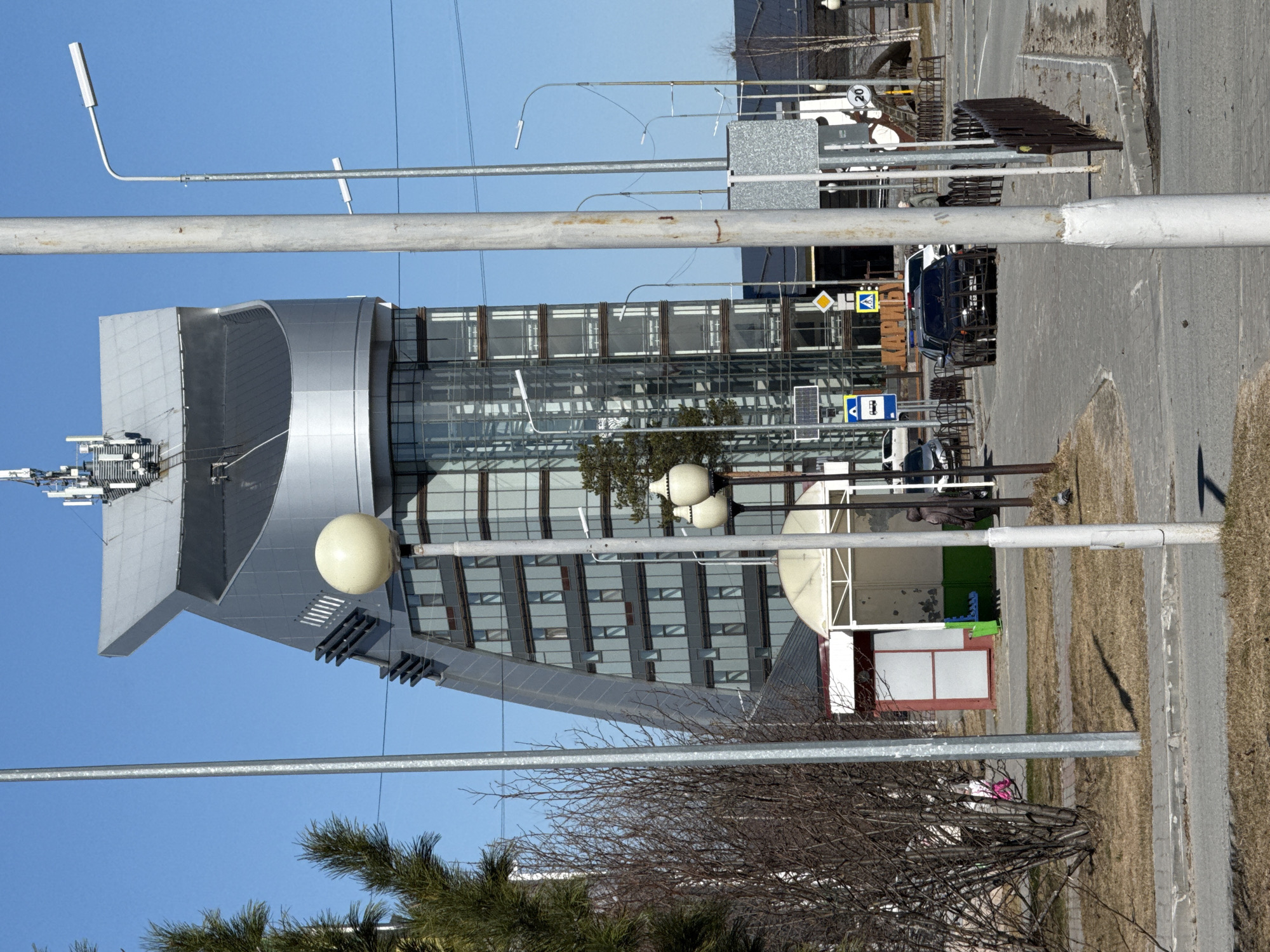 The image shows a modern building in Beloyarsky, Russia, characterized by a combination of silver metal cladding and extensive glass windows. The building has a distinctive, angular design with a notable section that curves. Various antennas and equipment are visible on the building's roof. Street lights are placed in front of the building, obscuring parts of it.
In the foreground, there are trees with bare branches, as well as evergreen trees, indicating a cool climate or early spring. A pedestrian walkway, a bus stop, a parking area, and a road are visible next to the building. A white van and a black vehicle are parked. In the distance, there are indistinct structures and landscape. The sky is clear and blue.