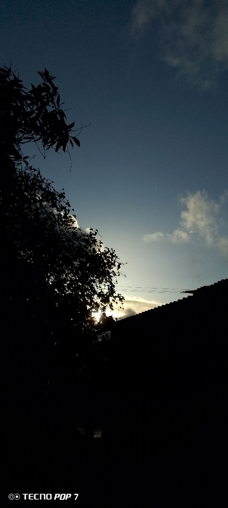 The image depicts a view towards the sky in Altagracia, Venezuela, taken during what appears to be dawn or dusk. The sky is a mix of blue and white, with clouds scattered around. Silhouetted in the foreground is foliage from trees on the left and the tiled roof of a building on the right. The sun is attempting to peek through the trees, casting a bright light behind the foliage. A couple of wires run across the scene, above the roof. The bottom of the image displays the text "Tecno Pop 7".
